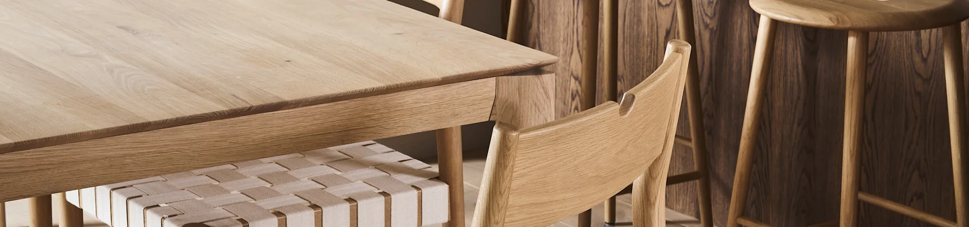 Gros plan d'une table à manger en bois clair avec une chaise à assise tressée et des tabourets en bois à l'arrière-plan.