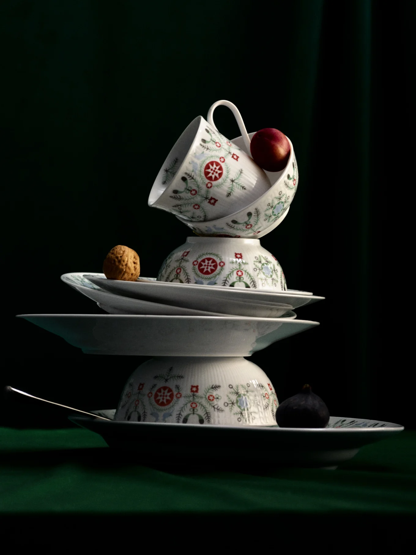Nature morte de vaisselle blanche empilée aux motifs festifs, avec une pomme, une noix, une figue et une cuillère sur fond vert foncé.