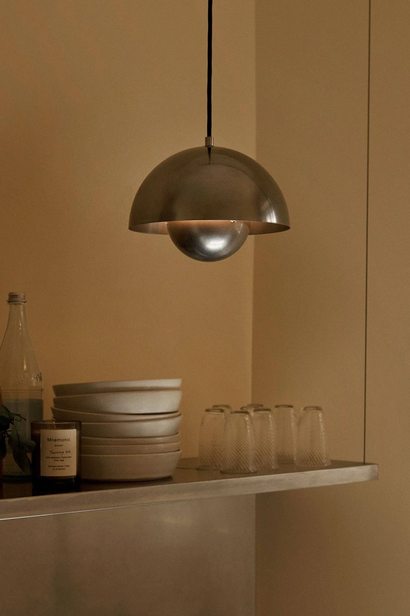 Suspension dôme métallique éclairant une étagère avec des bols empilés, des verres, une bouteille et une bougie.