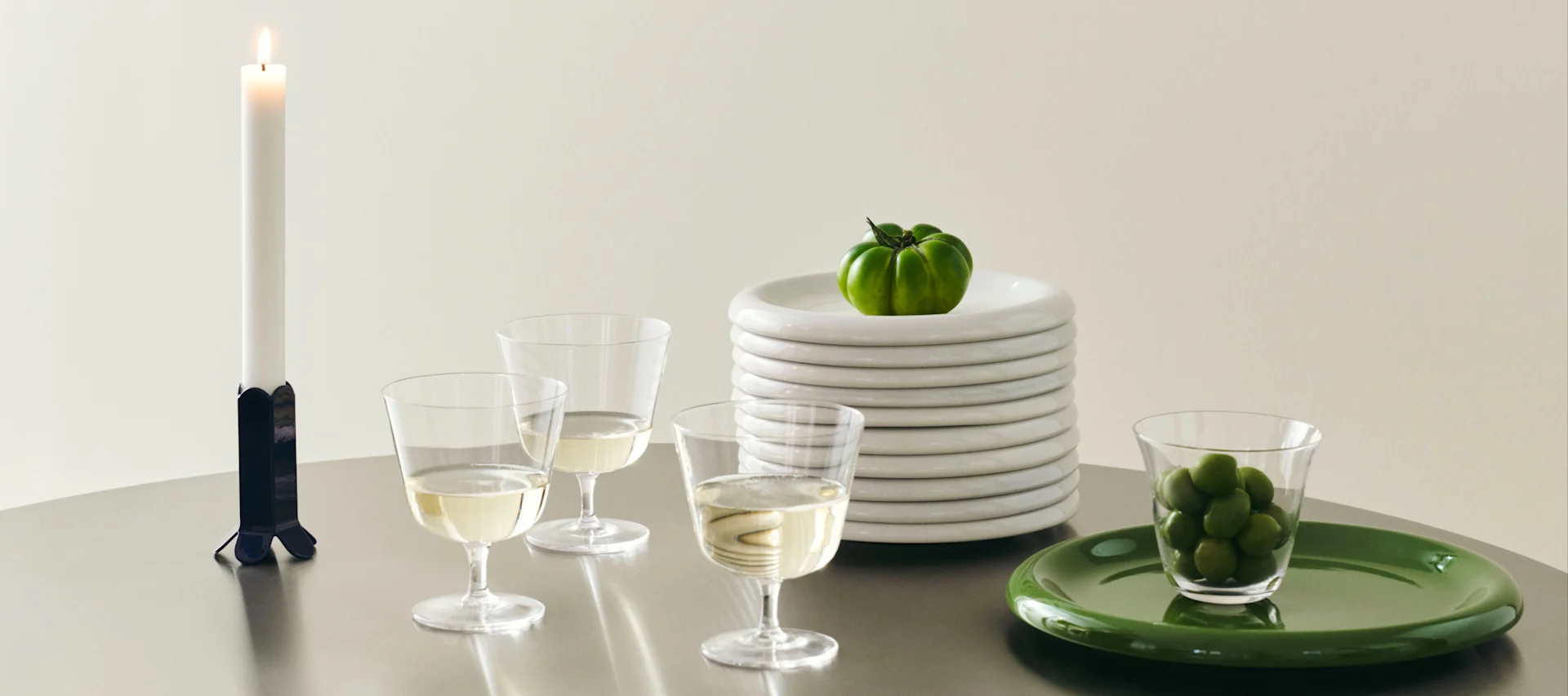 Table dressée avec bougie, verres à vin, pile d'assiettes blanches avec tomate verte, et olives vertes sur assiette verte.