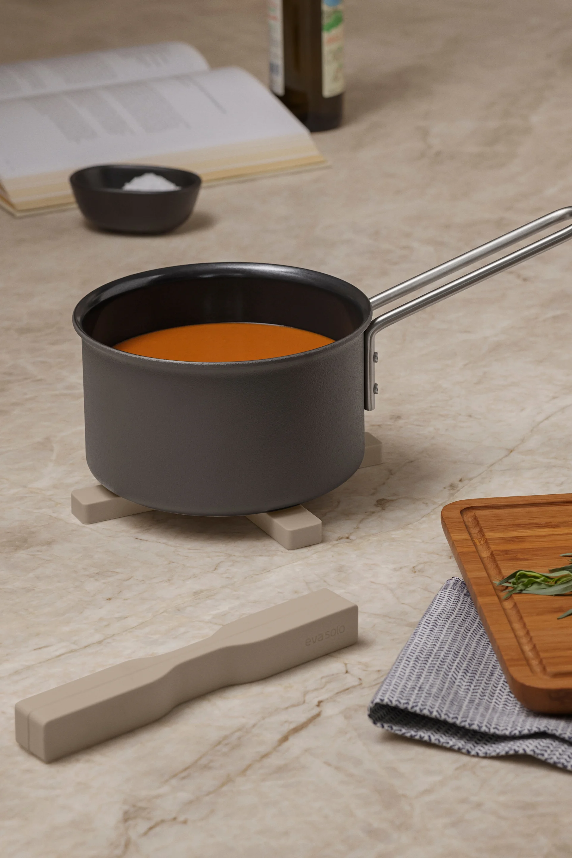 Casserole avec liquide orange sur un dessous-de-plat, avec livre de cuisine, sel, fouet, herbes sur planche et ustensile sur comptoir en marbre.