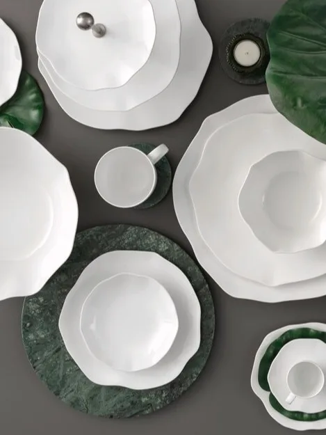 Une collection de vaisselle en céramique blanche aux bords ondulés, disposée avec des feuilles vertes et des dessous de verre en marbre sur une surface sombre.