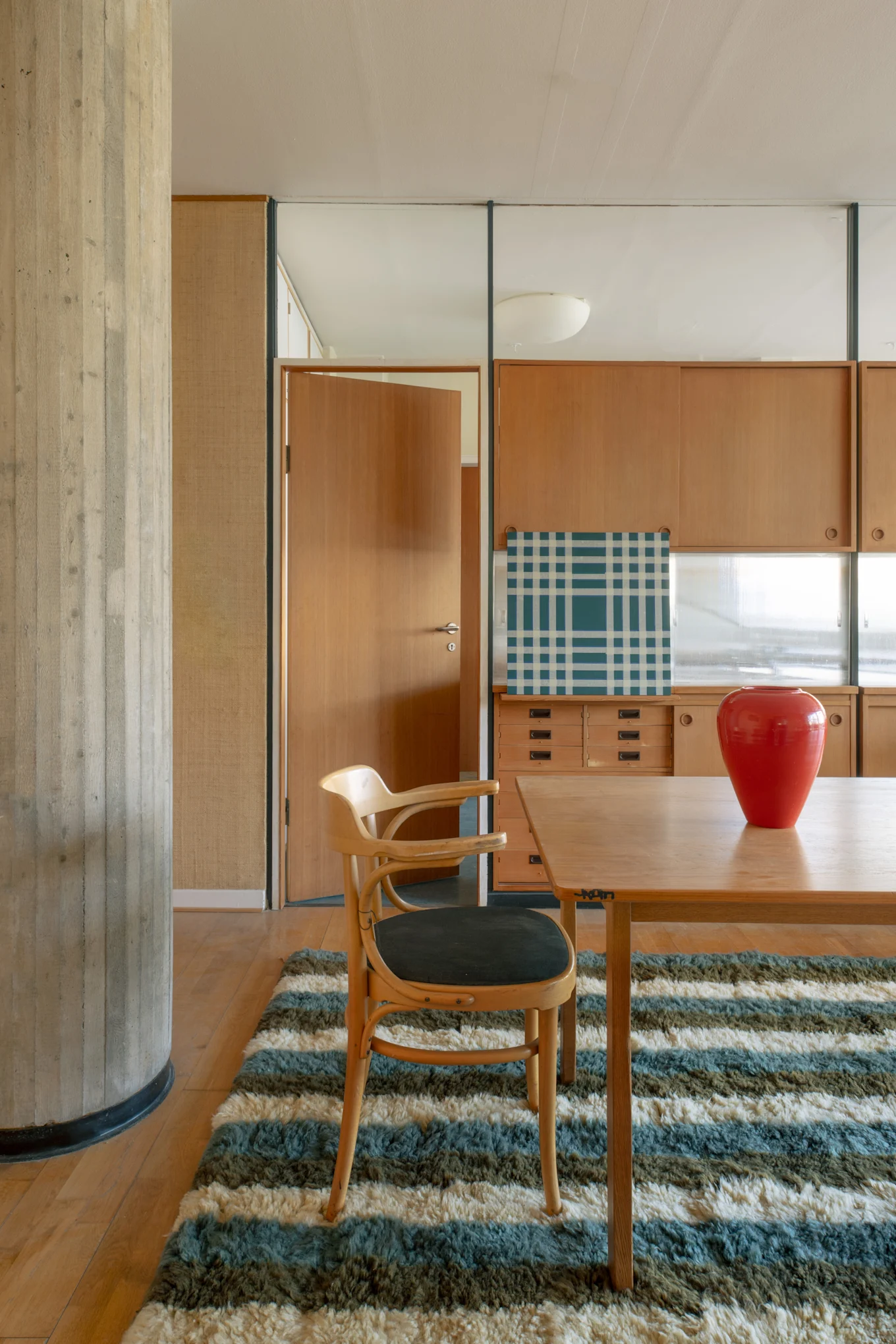 Intérieur moderne avec une table à manger en bois, une chaise, un vase rouge et un tapis rayé. Un pilier en béton encadre une armoire en bois et une porte ouverte.