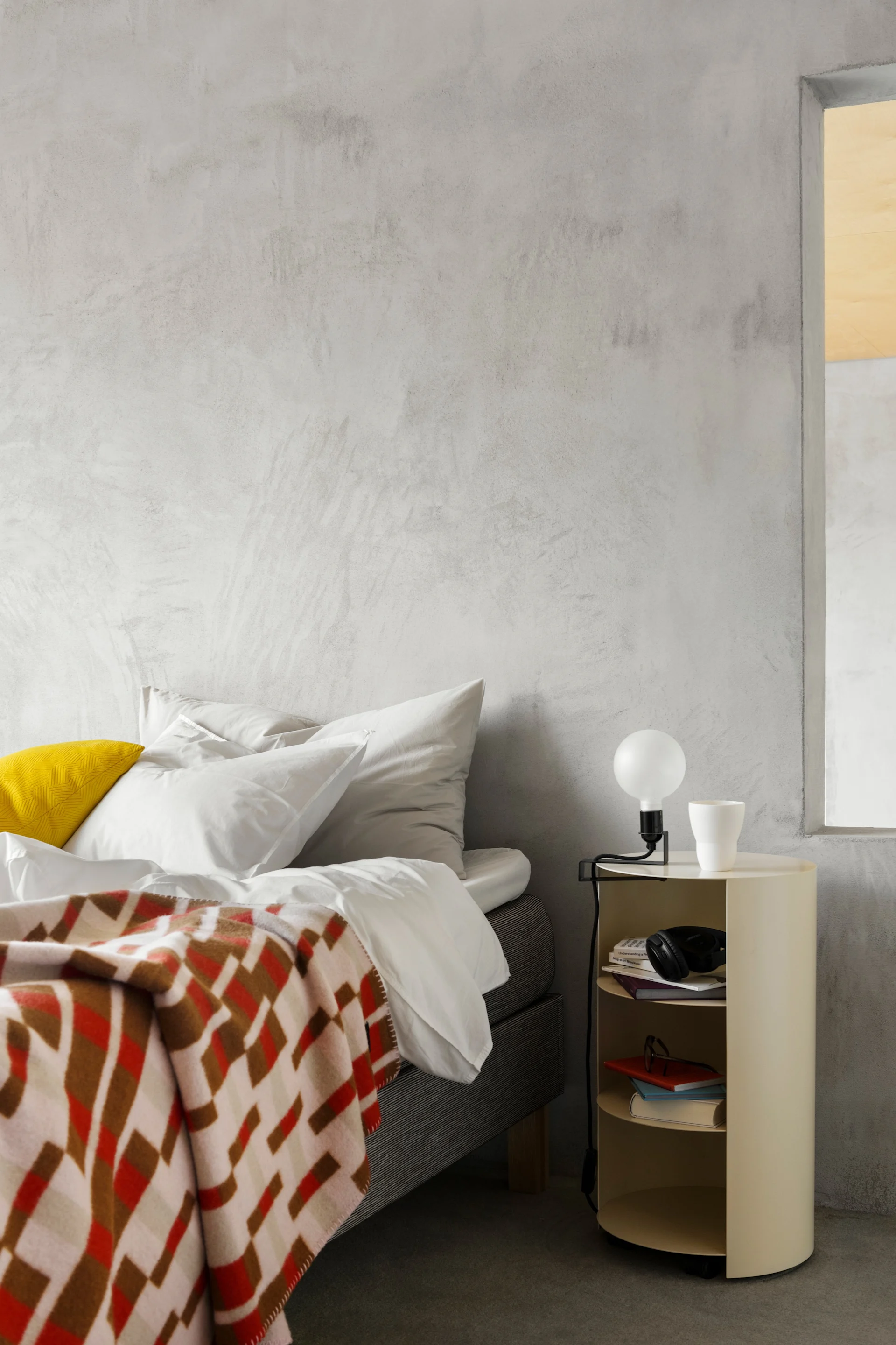 Chambre à coucher moderne avec un lit, une couverture à motifs, un coussin jaune et une table de chevet couleur crème sur laquelle sont posés une lampe et des livres.
