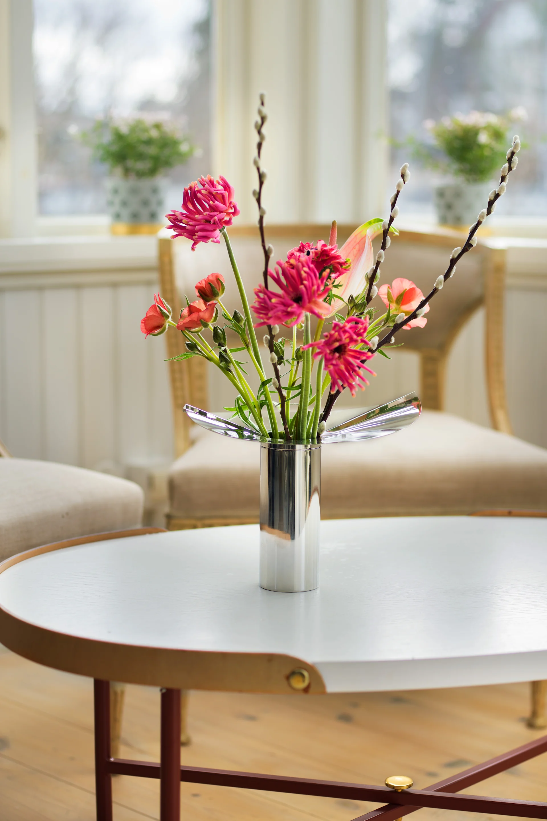 Une table basse ronde est décorée d'un vase/bougeoir Löv chromé de Klong, dans lequel sont disposées des fleurs printanières.