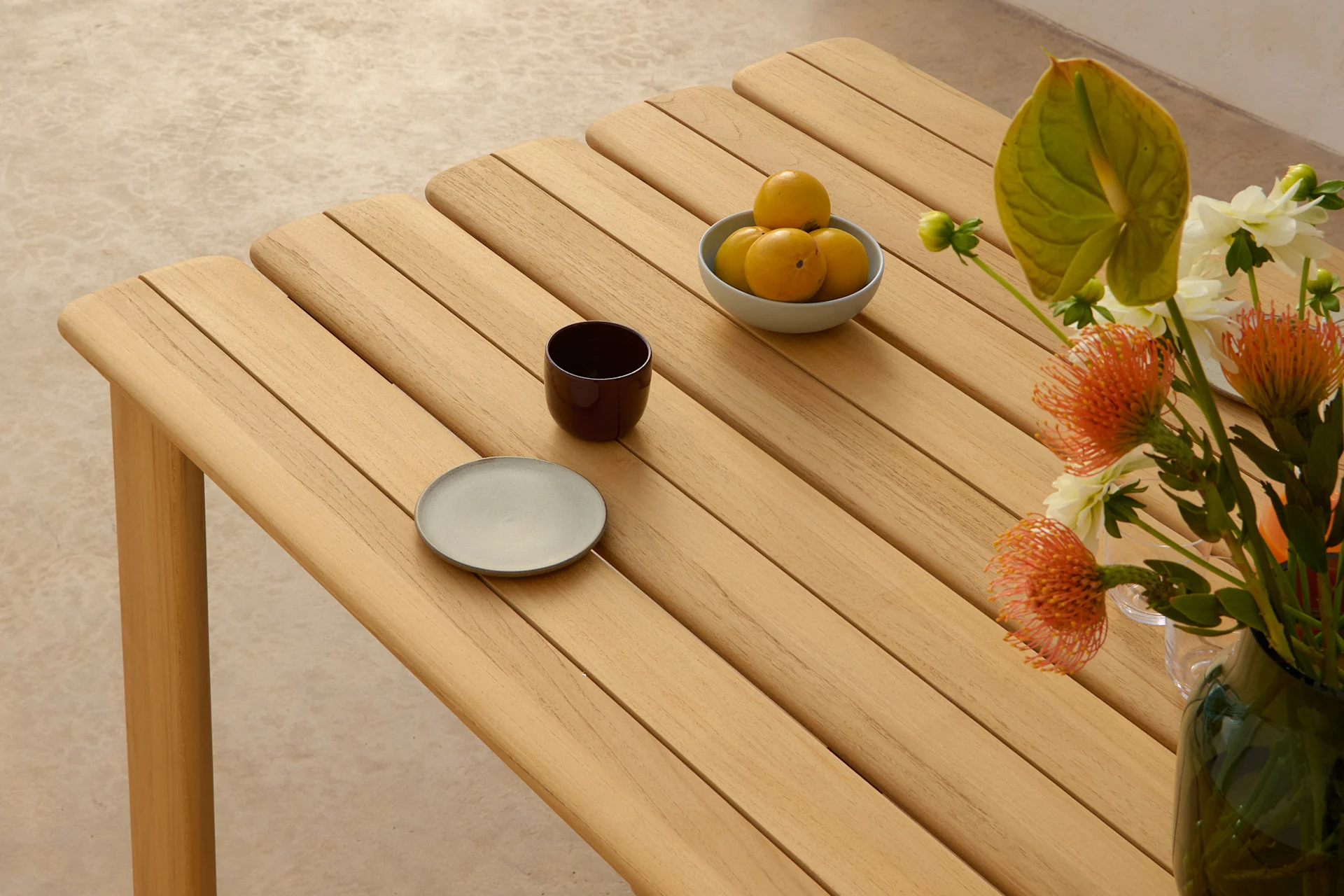 Une table à lattes en bois clair avec un bol de fruits jaunes, une tasse foncée, une assiette grise et un vase de fleurs oranges et blanches.