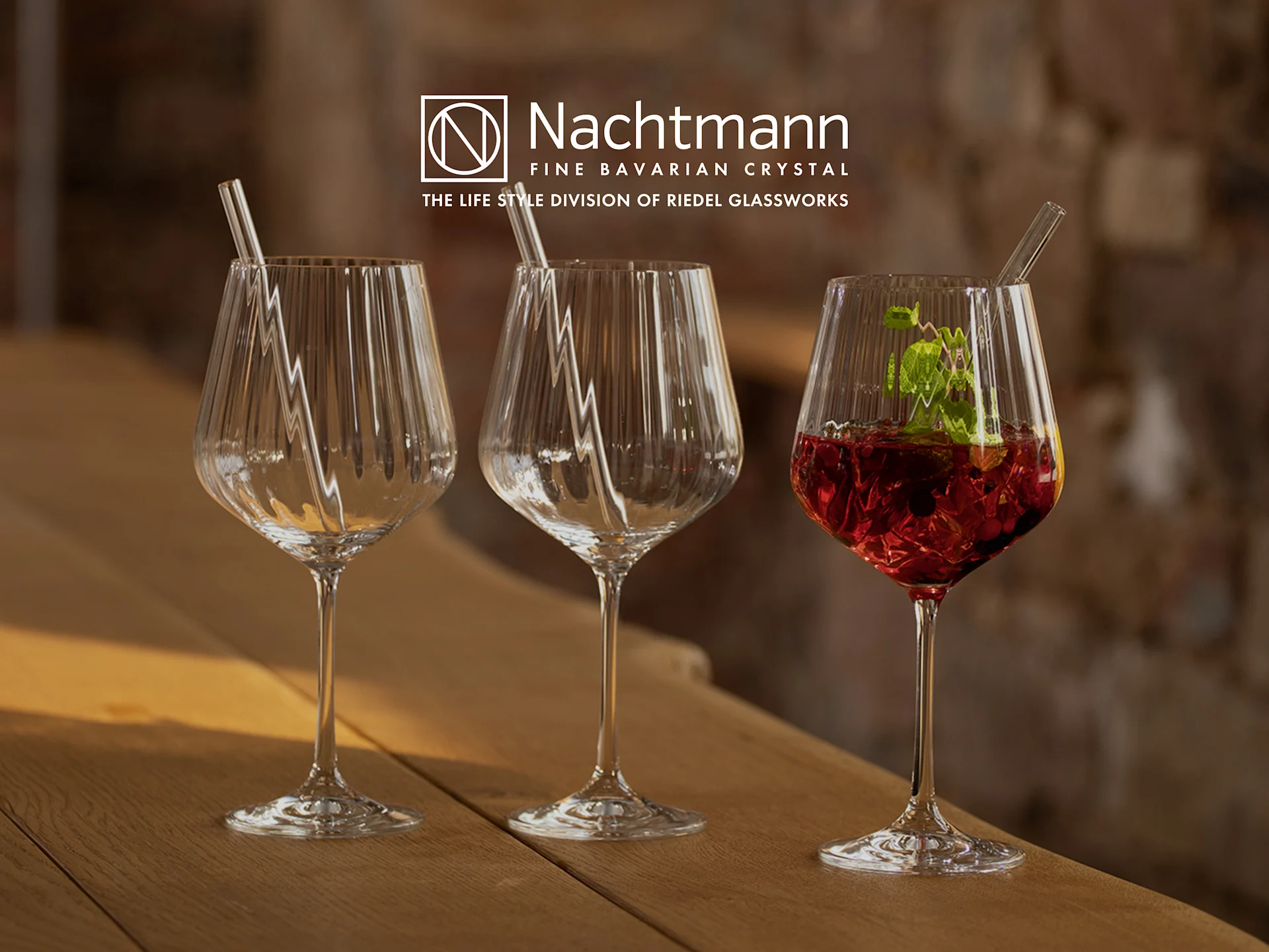 Trois verres en cristal Nachtmann sur une table en bois, deux vides avec des pailles, un avec une boisson rouge et de la menthe.