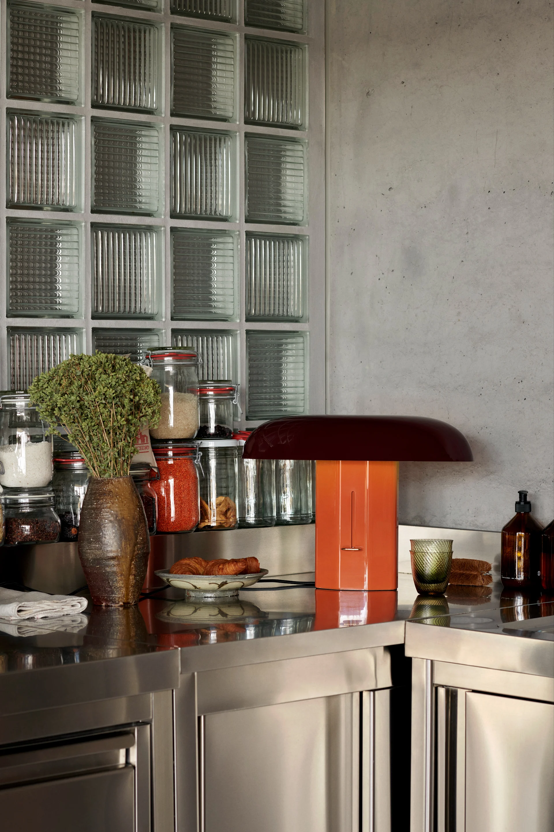 Un comptoir de cuisine en acier inoxydable avec un vase brun d'herbes, des bocaux d'ingrédients, une lampe orange en forme de champignon, des croissants et des bouteilles en verre.
