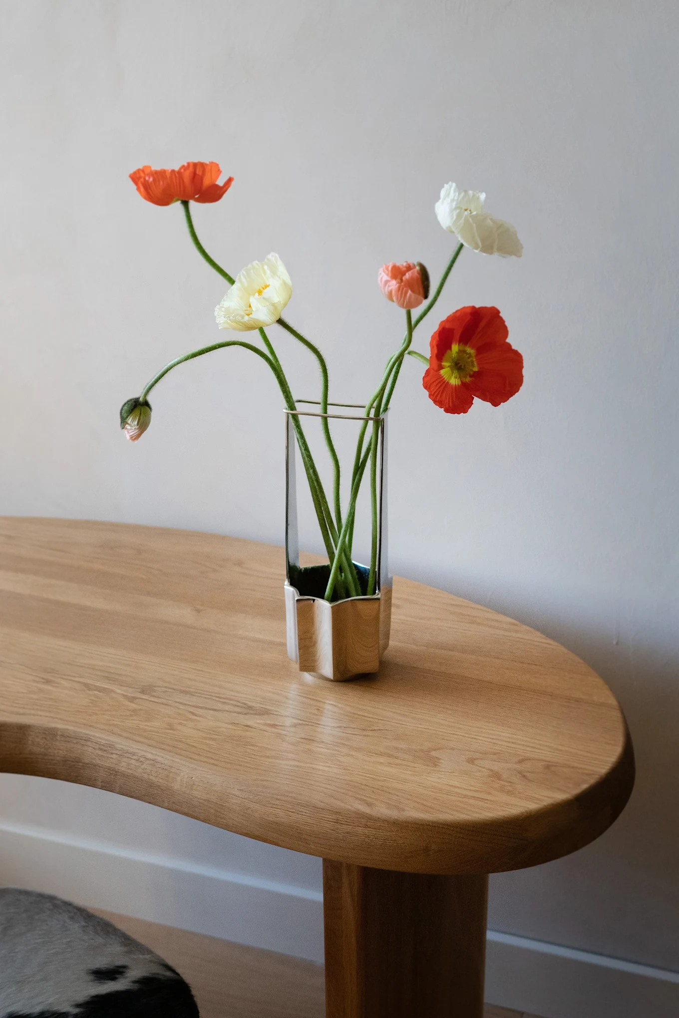 Un bouquet de coquelicots oranges, jaunes et blancs avec de longues tiges vertes dans un vase unique en verre et argent sur une table en bois clair.