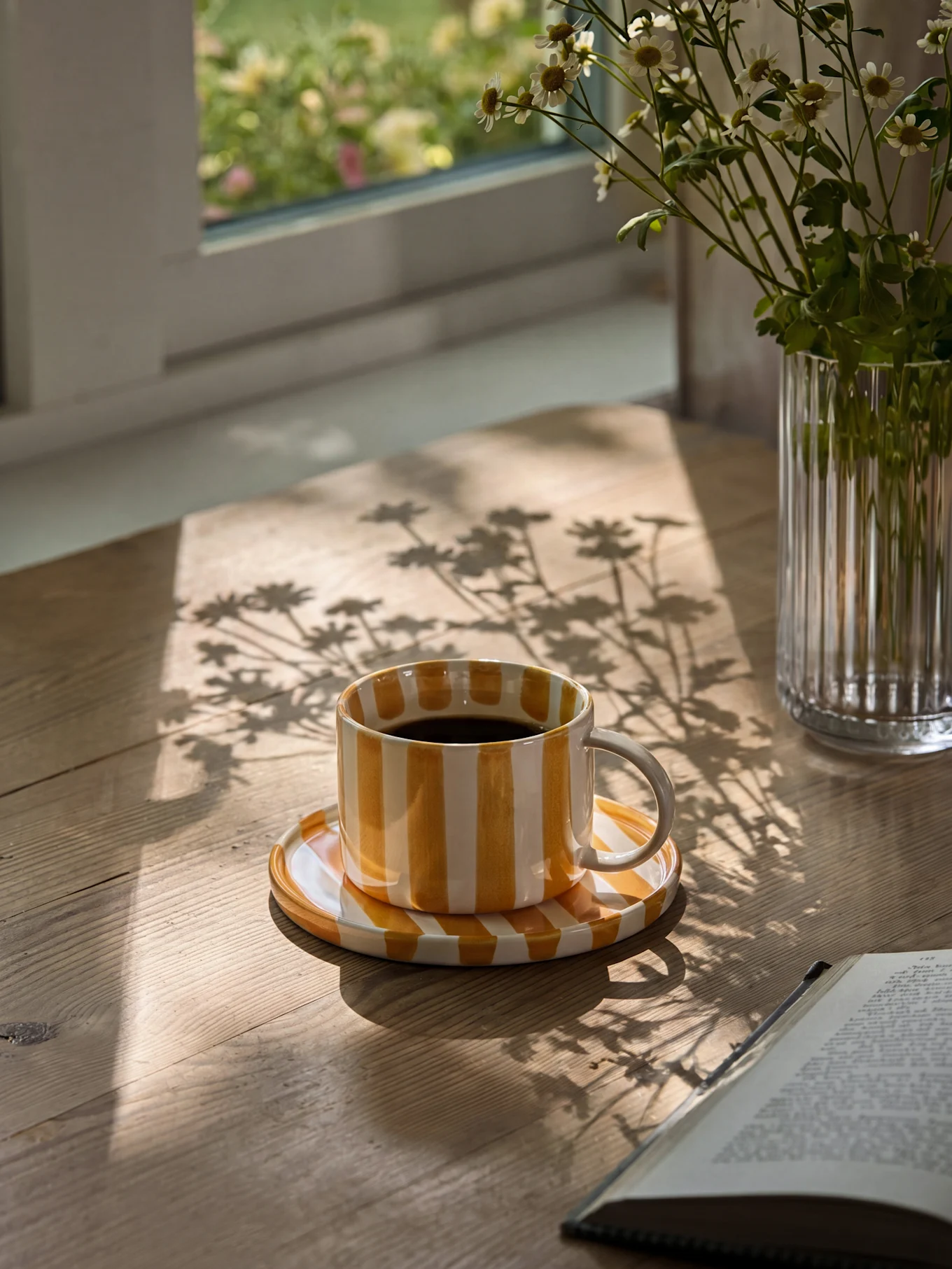 Tasse à café rayée jaune et blanche avec du café noir sur une table en bois avec des ombres de soleil et un vase de marguerites.