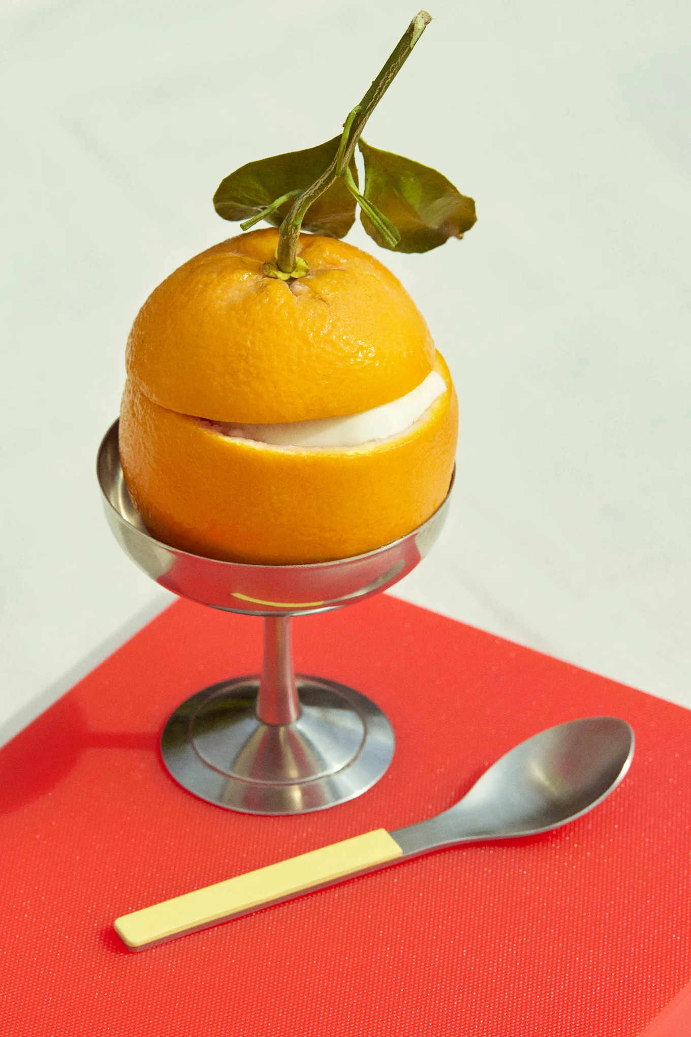 Dessert à l'orange avec garniture blanche dans une coupe argentée, avec une cuillère, sur une surface rouge.
