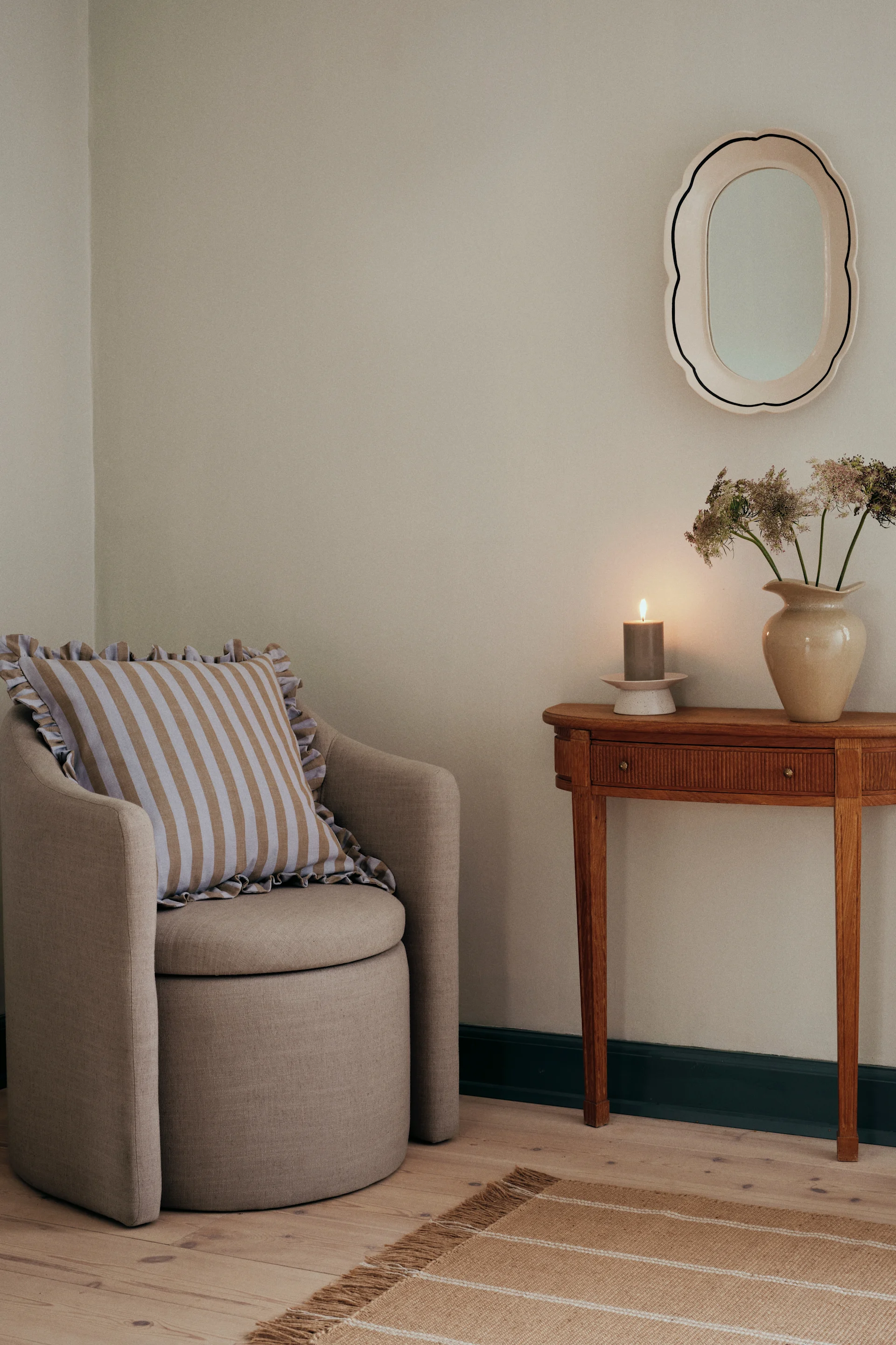 Un fauteuil avec un coussin rayé, une table d'appoint en bois avec une bougie allumée et un vase de fleurs, un miroir et un tapis rayé.