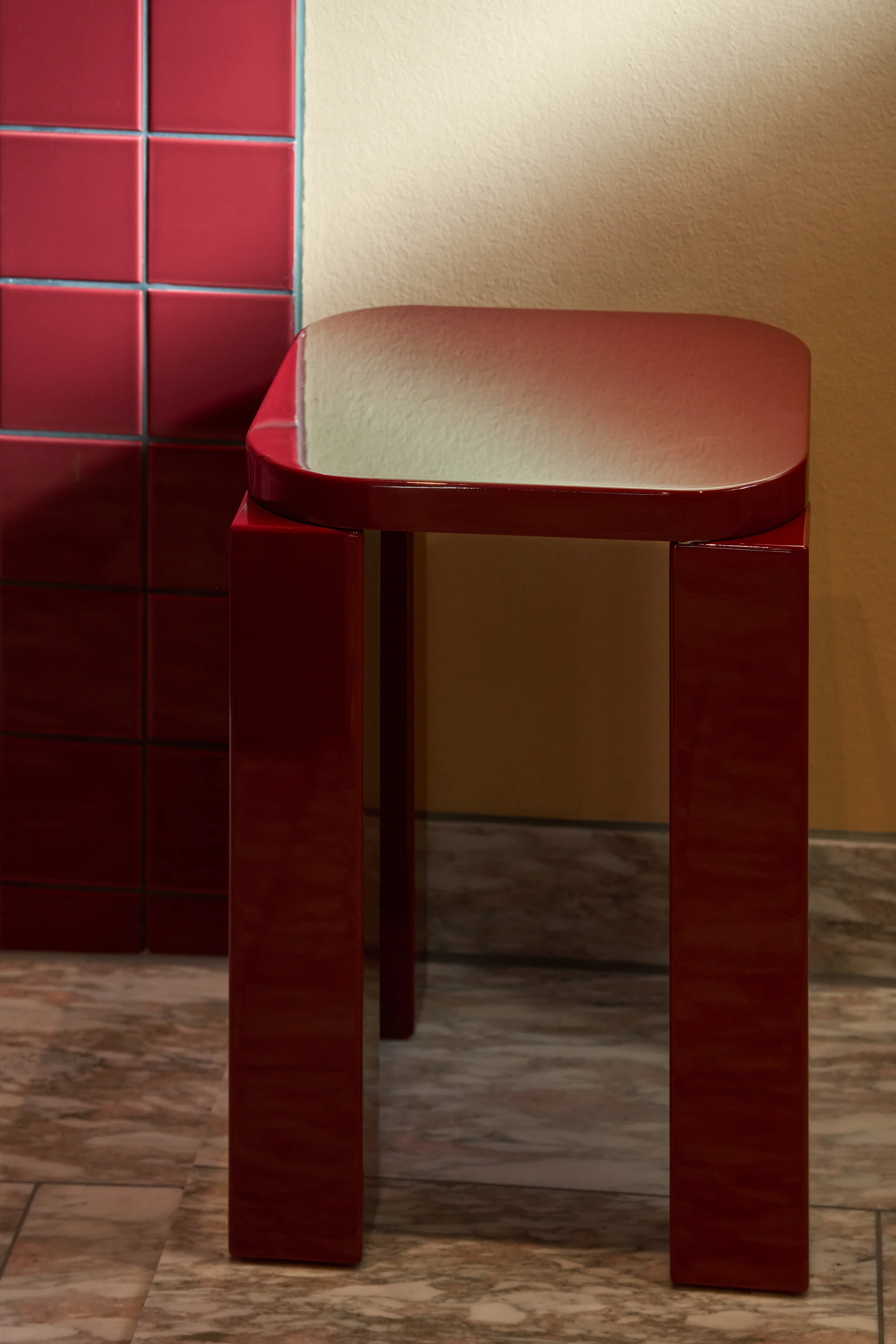Un tabouret rouge foncé brillant avec un plateau rectangulaire et quatre pieds repose sur un sol en marbre à côté d'un mur carrelé.