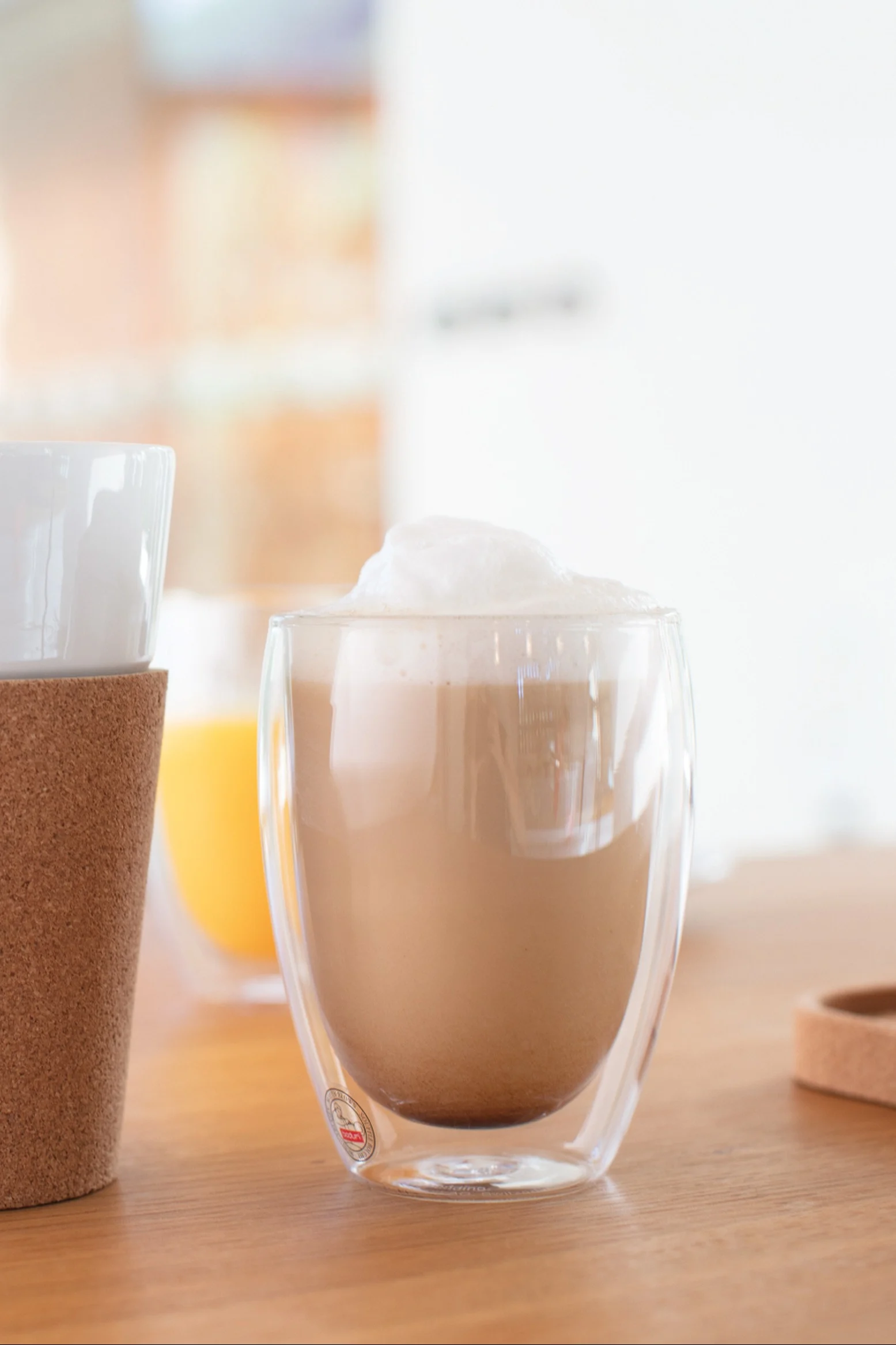 Latte mousseux dans une tasse en verre transparent à double paroi sur une table en bois, avec une autre tasse avec manchon en liège à gauche et une boisson orange en arrière-plan.