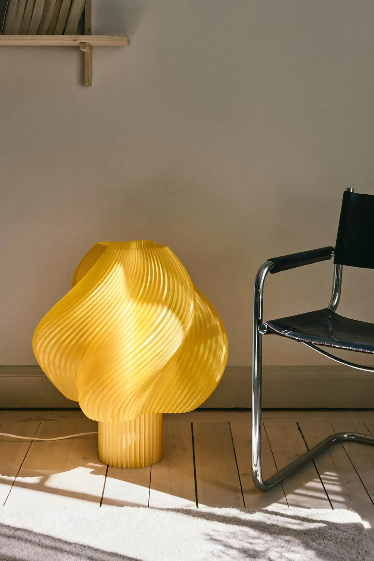 Une lampe de table jaune vif et sculpturale au design côtelé et ondulé brille sur un parquet à côté d'une chaise en cuir noir et chrome.