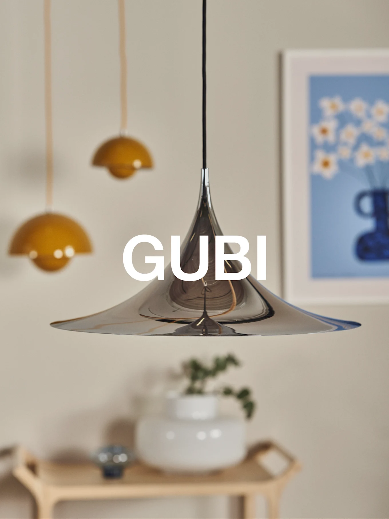 Une lampe suspendue conique en chrome poli avec le logo GUBI, sur un fond flou avec d'autres lampes et de l'art.