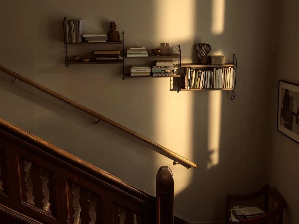 Mur ensoleillé avec étagères en bois remplies de livres et de décorations, à côté d'un escalier et d'une rampe en bois.