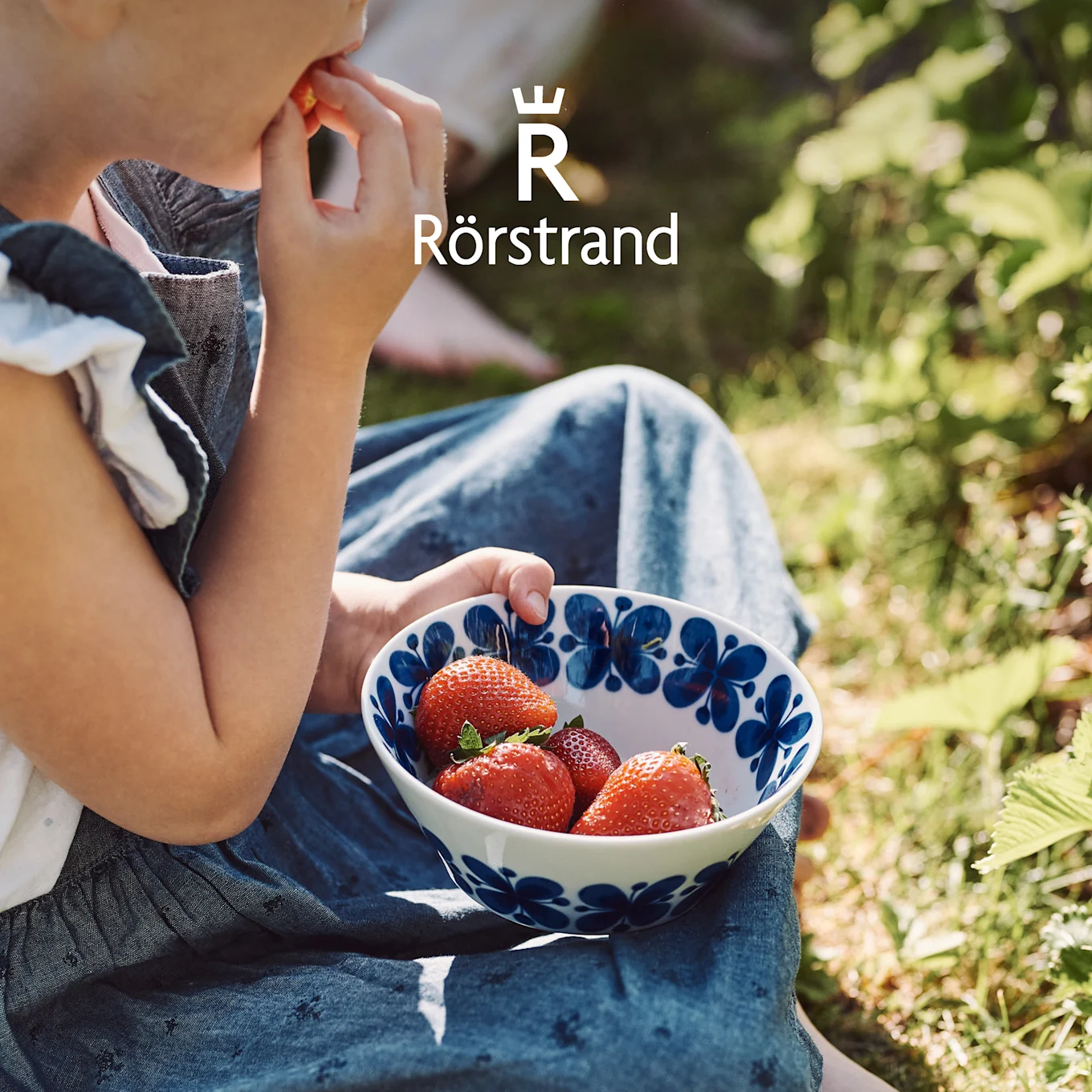Un enfant mange des fraises dans un bol Rörstrand blanc aux motifs floraux bleus, assis dans un jardin ensoleillé.