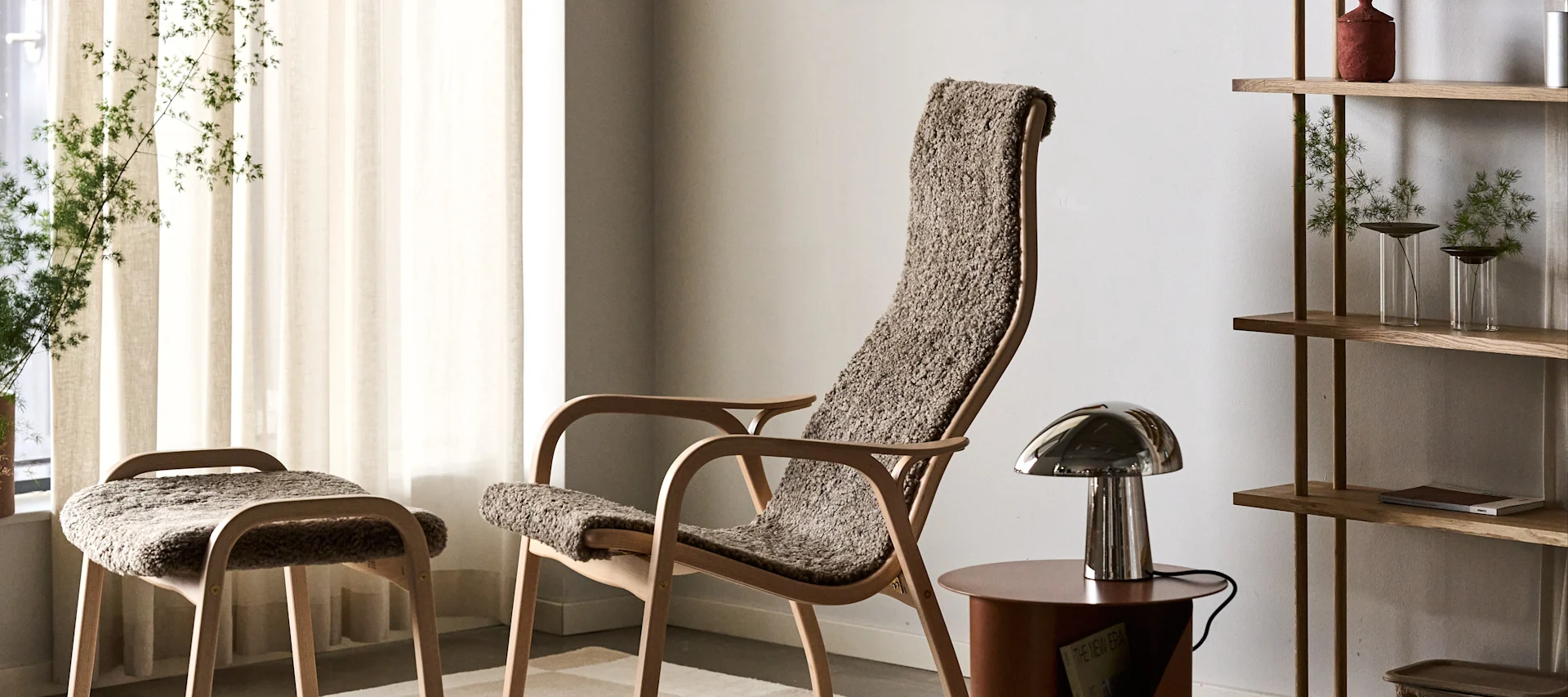 Fauteuil et tabouret en bois clair avec revêtement brun texturé, lampe de table métallique et étagère en bois avec plantes.