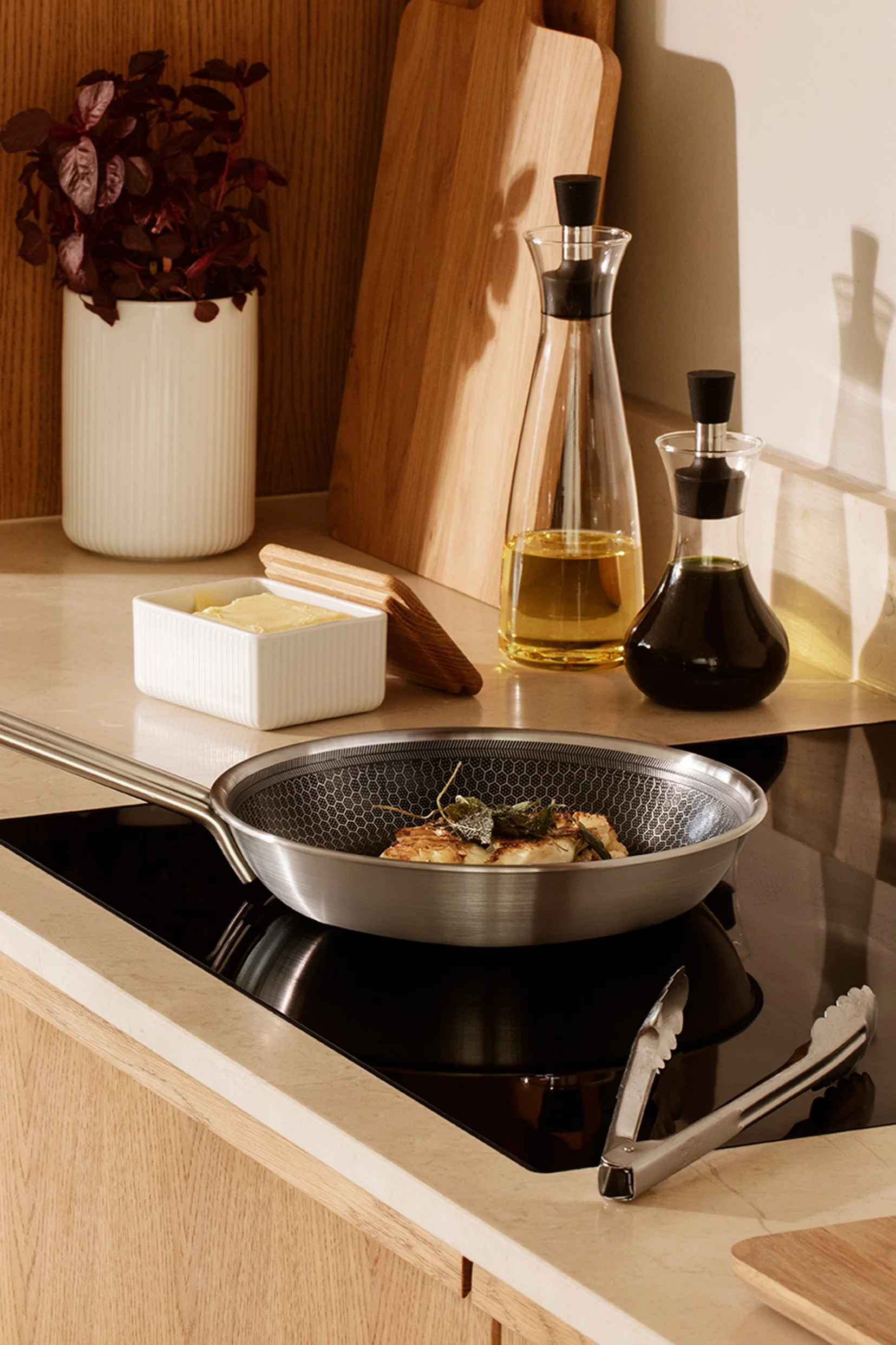 Poêle en acier inoxydable avec aliments sur plaque à induction, à côté de beurre, bouteilles d'huile et ustensiles de cuisine.