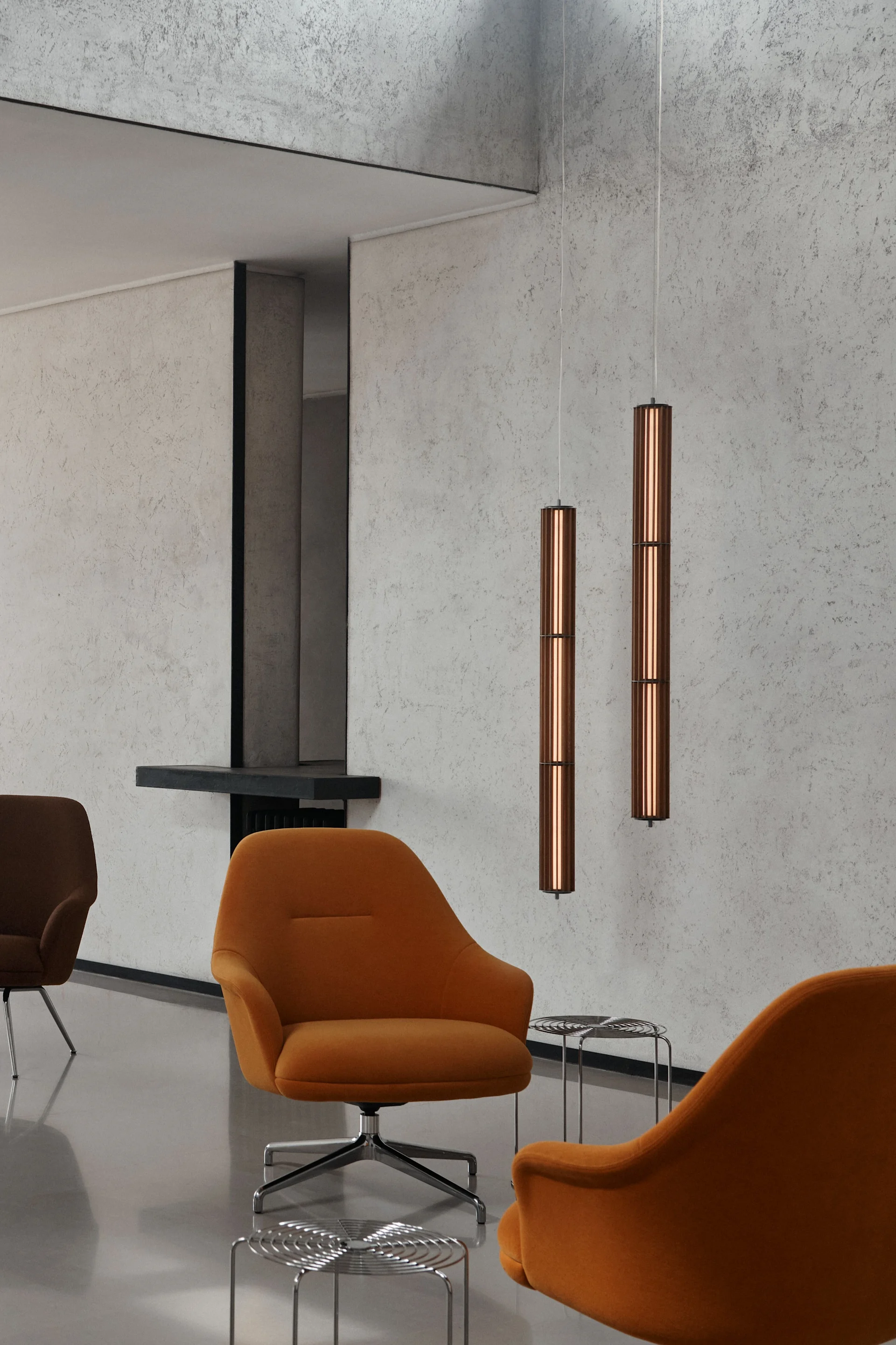 Deux suspensions cylindriques en cuivre dans une pièce moderne avec des fauteuils orange et des murs texturés en béton.