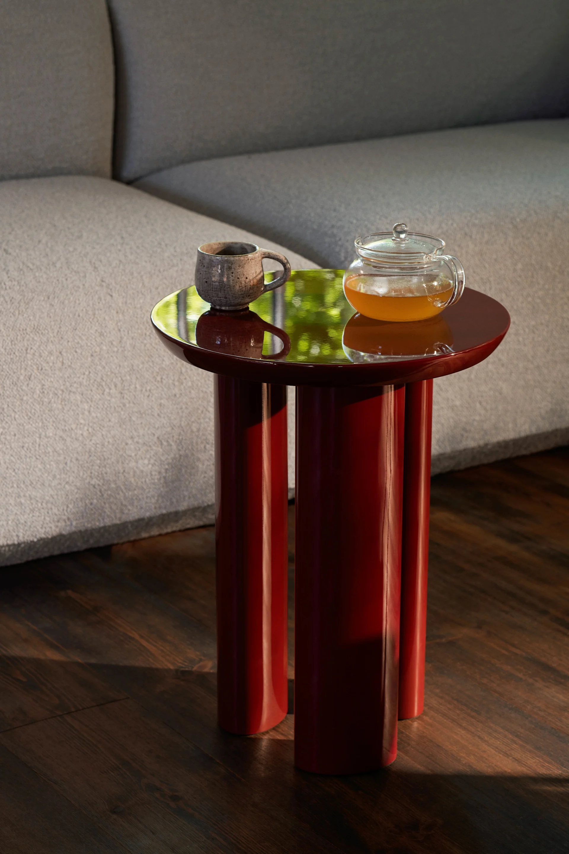 Table d'appoint rouge brillante à trois pieds, sur laquelle sont posés une tasse en céramique et une théière en verre remplie de thé ambré.