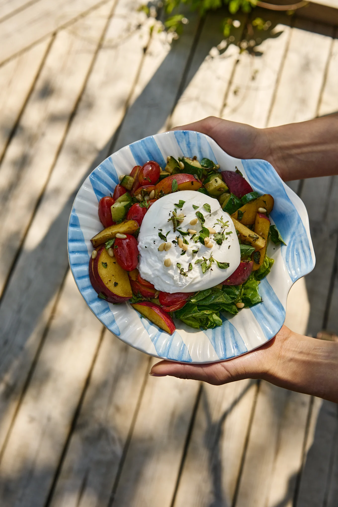 Mains tenant une salade de burrata avec tomates, pêches, concombre et légumes verts dans un bol rayé en forme de coquille.
