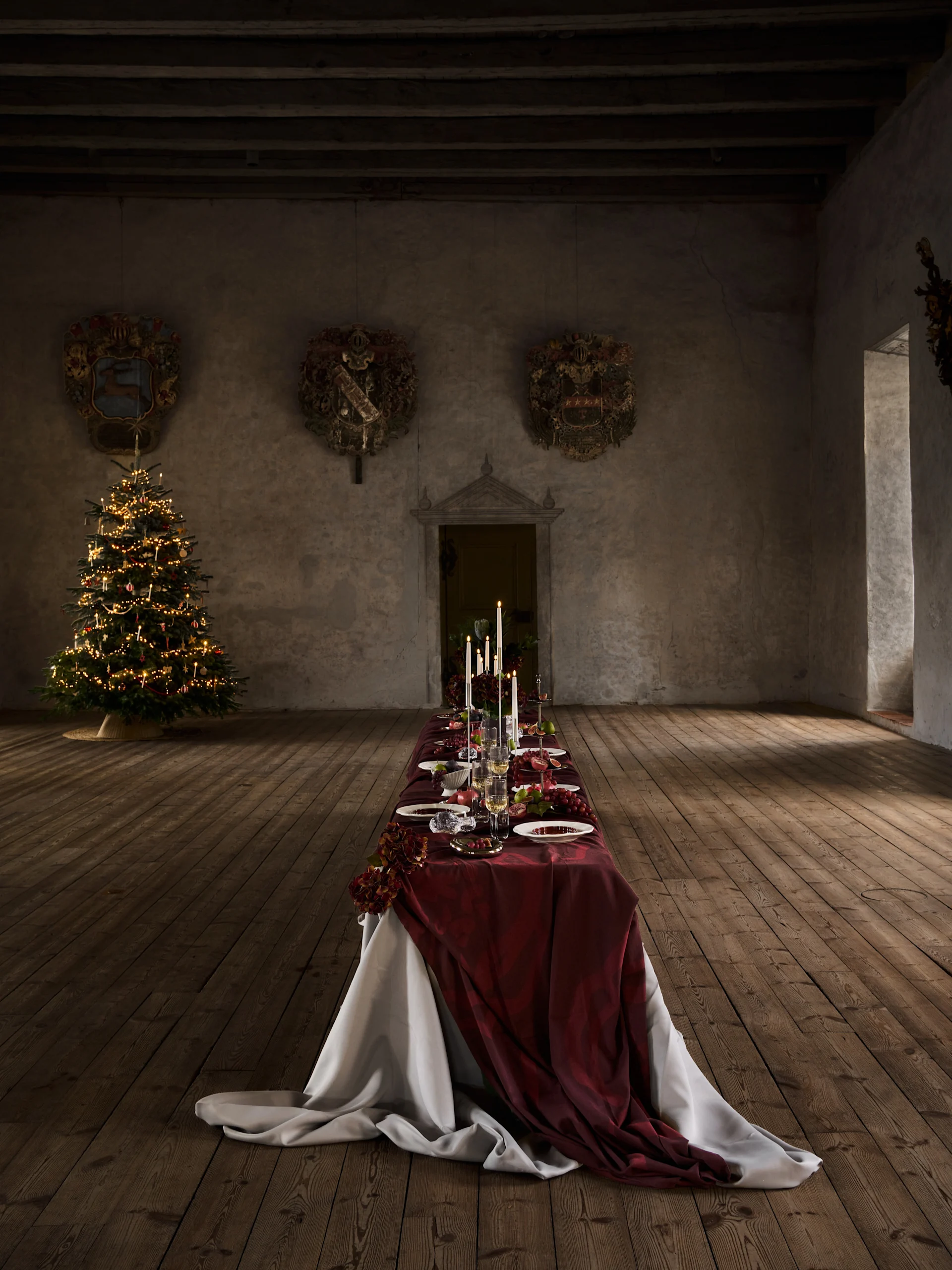 Longue table de dîner de Noël avec nappe rouge, bougies, fruits et couverts, à côté d'un sapin illuminé dans une vieille salle.