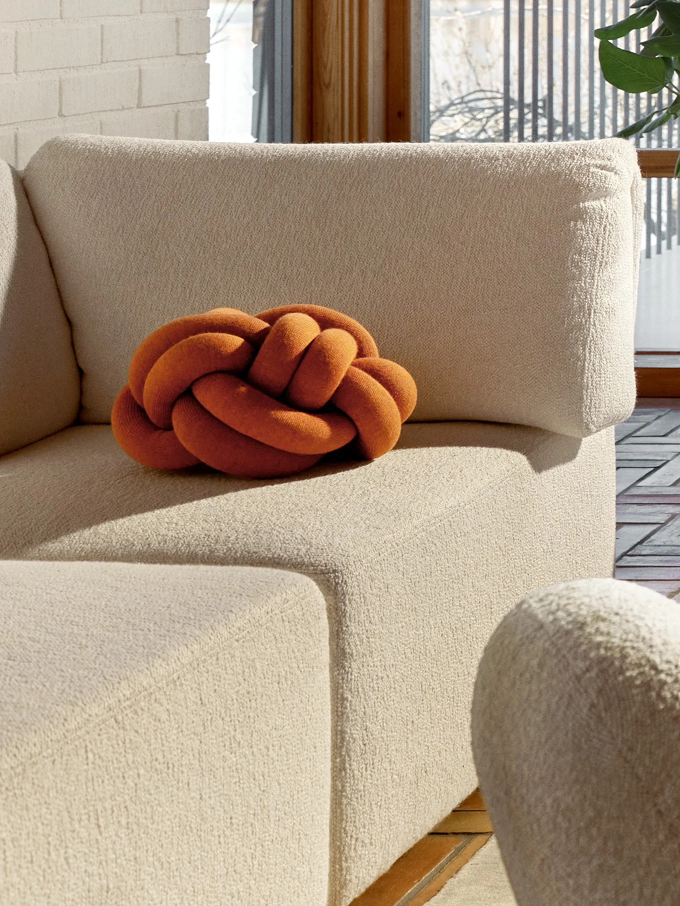 Un coussin orange en forme de nœud sur un canapé beige texturé, avec un mur de briques blanches et une fenêtre en arrière-plan.