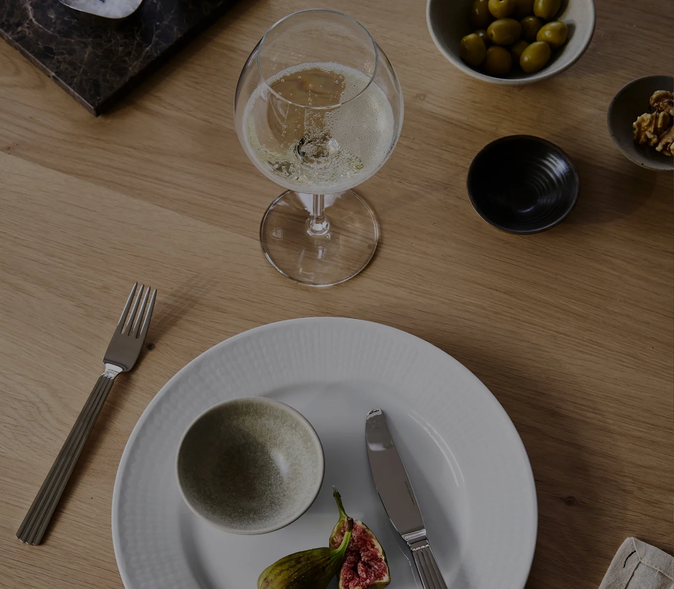 Un verre de champagne, une assiette blanche avec un bol et des figues, une fourchette, un couteau, des olives et des noix sur une table en bois.