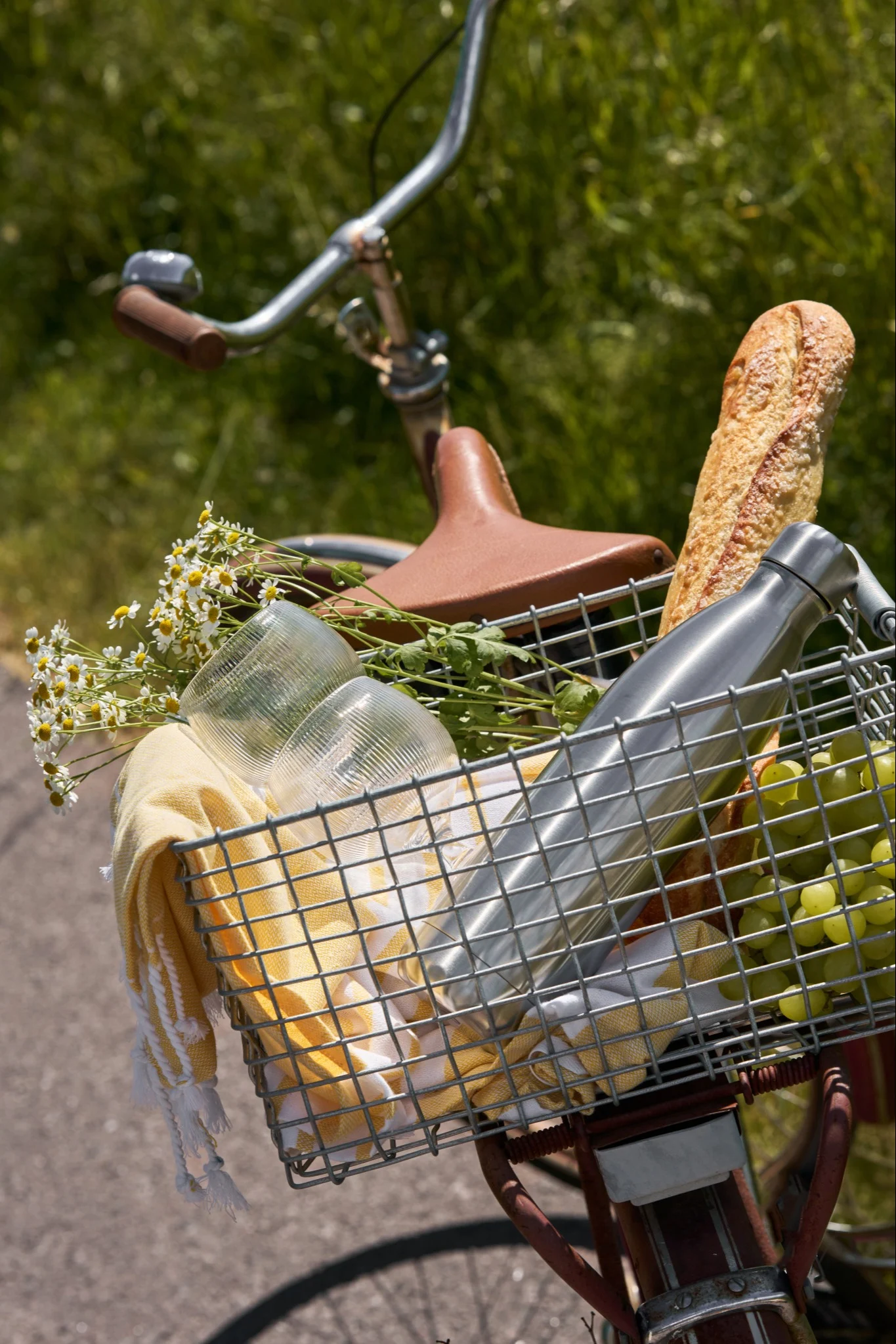 Vélo avec un panier avant rempli d'articles de pique-nique : pain, raisins verts, une bouteille d'eau argentée, des tasses, une serviette jaune et des fleurs.