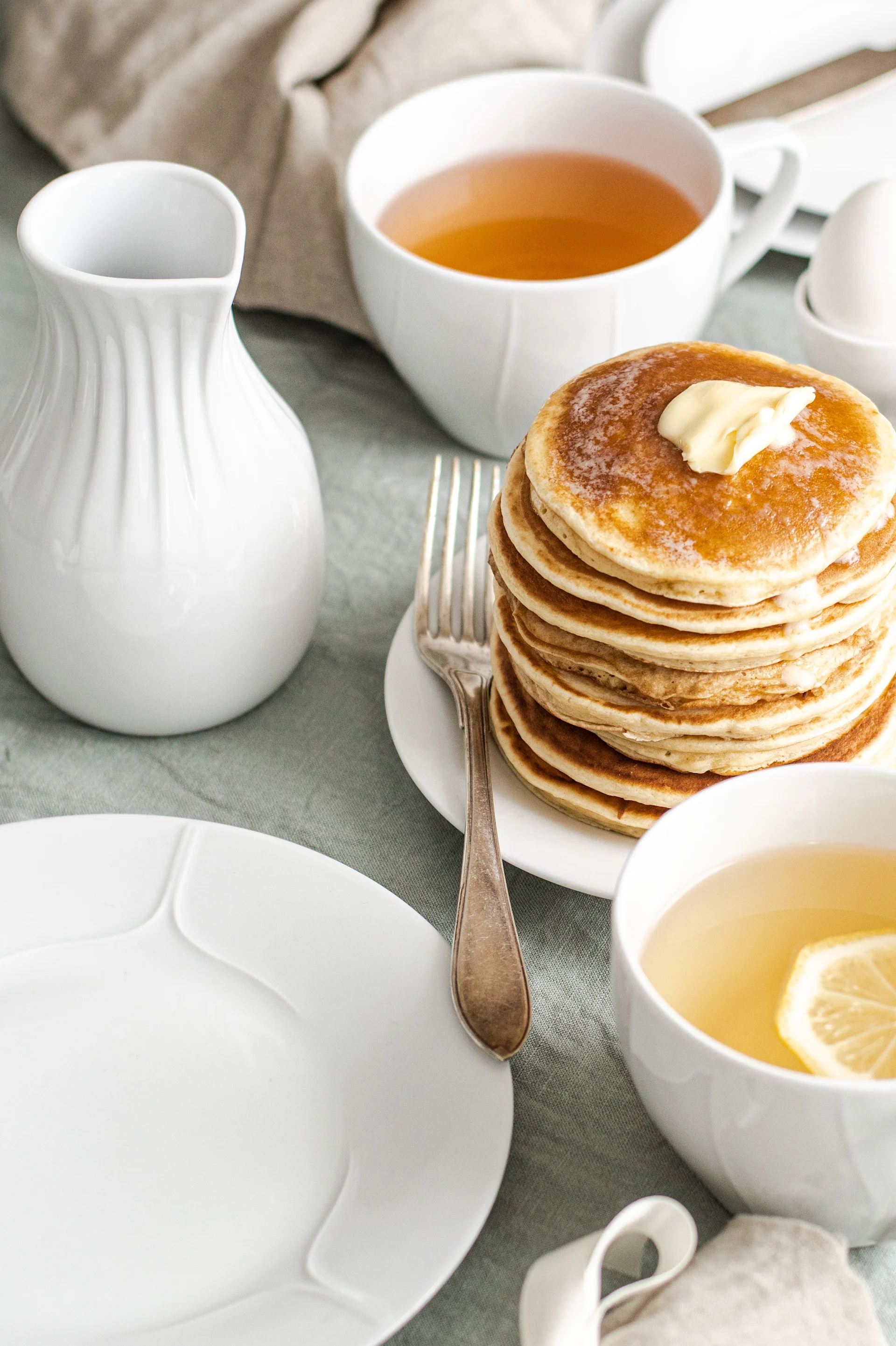 Pile de crêpes avec du beurre fondu, servies avec du thé et un pichet sur une nappe vert clair.