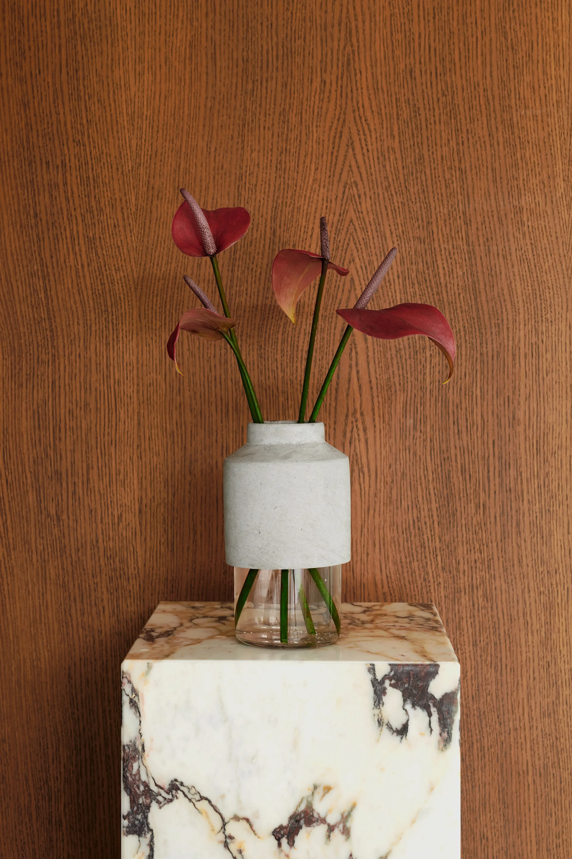 Des fleurs d'anthurium rouges dans un vase bicolore en verre et gris, posé sur un socle en marbre blanc devant un mur en bois.