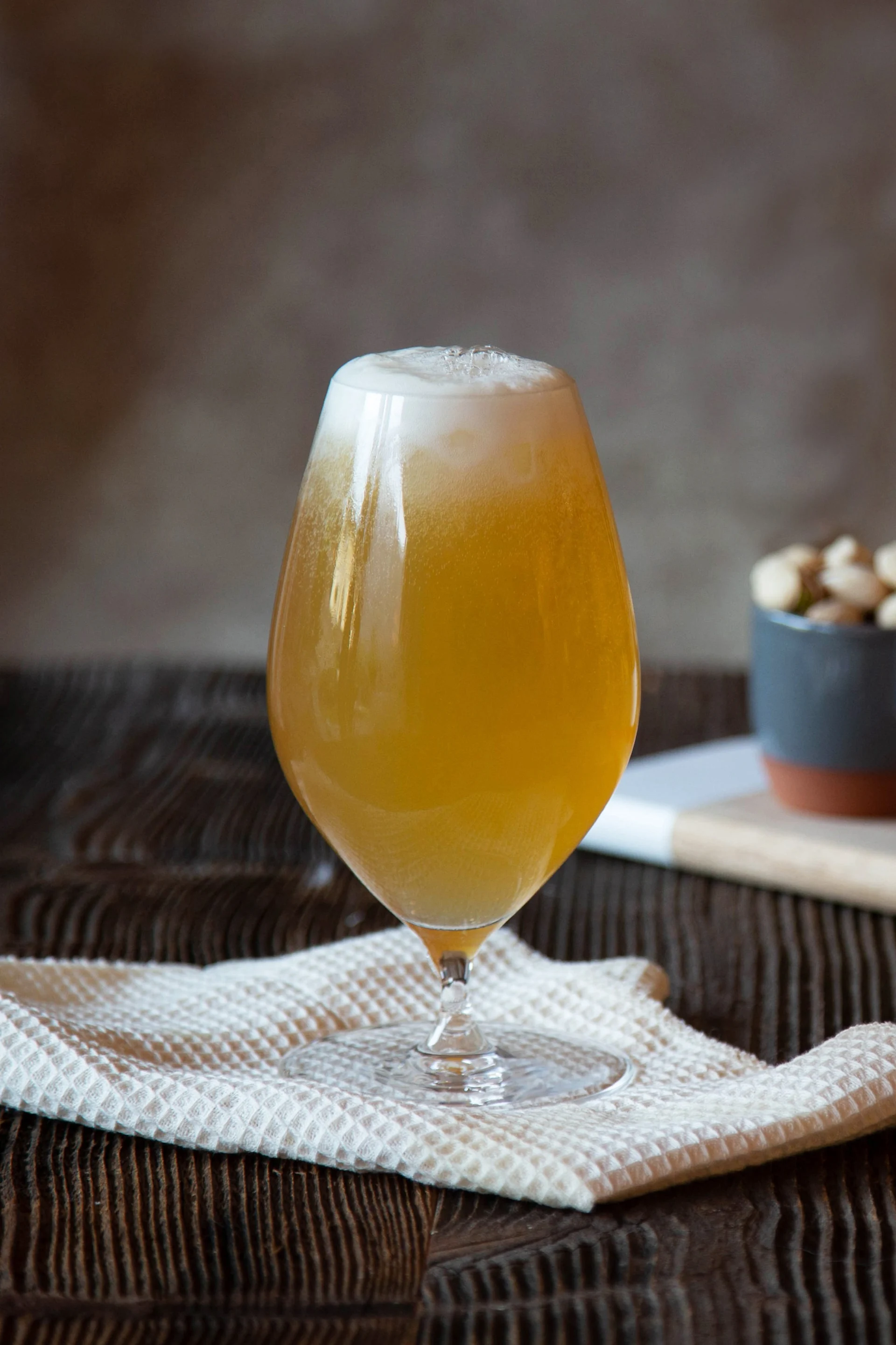 Un verre de bière ambrée mousseuse sur un tissu blanc, avec des bols de noix et de gressins à l'arrière-plan sur une planche en bois.