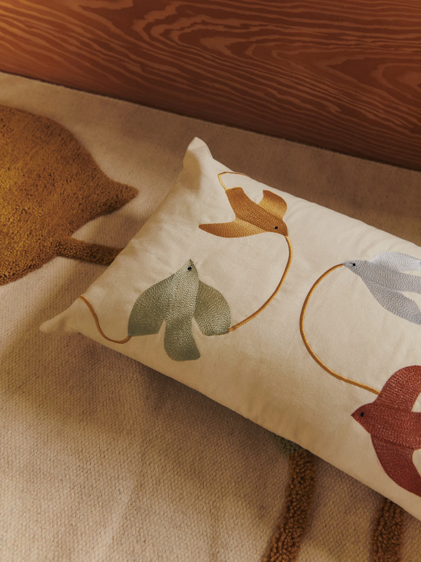 Coussin lombaire avec oiseaux et feuilles brodés or, vert, bleu et marron rougeâtre sur un tapis clair.