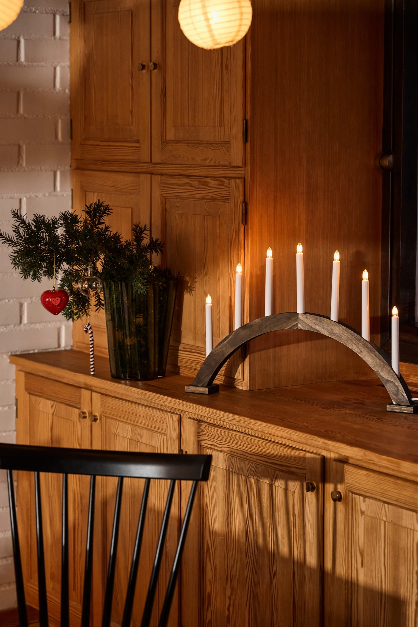 Un bougeoir de l'Avent en forme d'arc, en bois teinté foncé, est posé sur une commode en bois, à côté d'un vase rempli de branches de sapin.