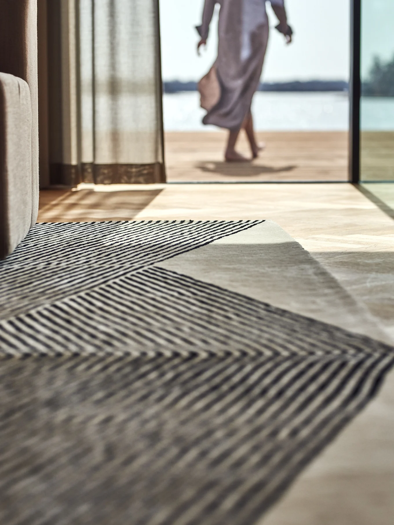 Tapis à rayures géométriques sur un parquet, avec une personne marchant vers un lac à l'extérieur.