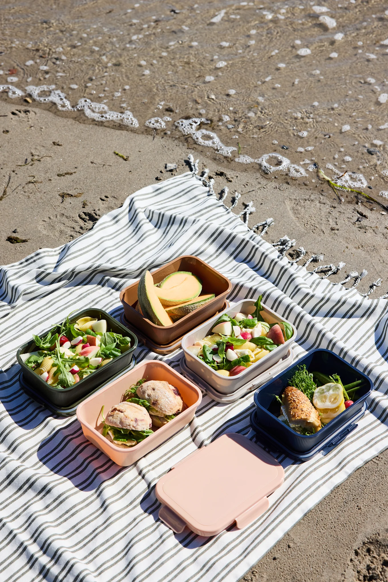 Cinq boîtes à lunch colorées ouvertes avec salades, sandwichs et melon, plus une boîte rose fermée, sur une couverture rayée à la plage.