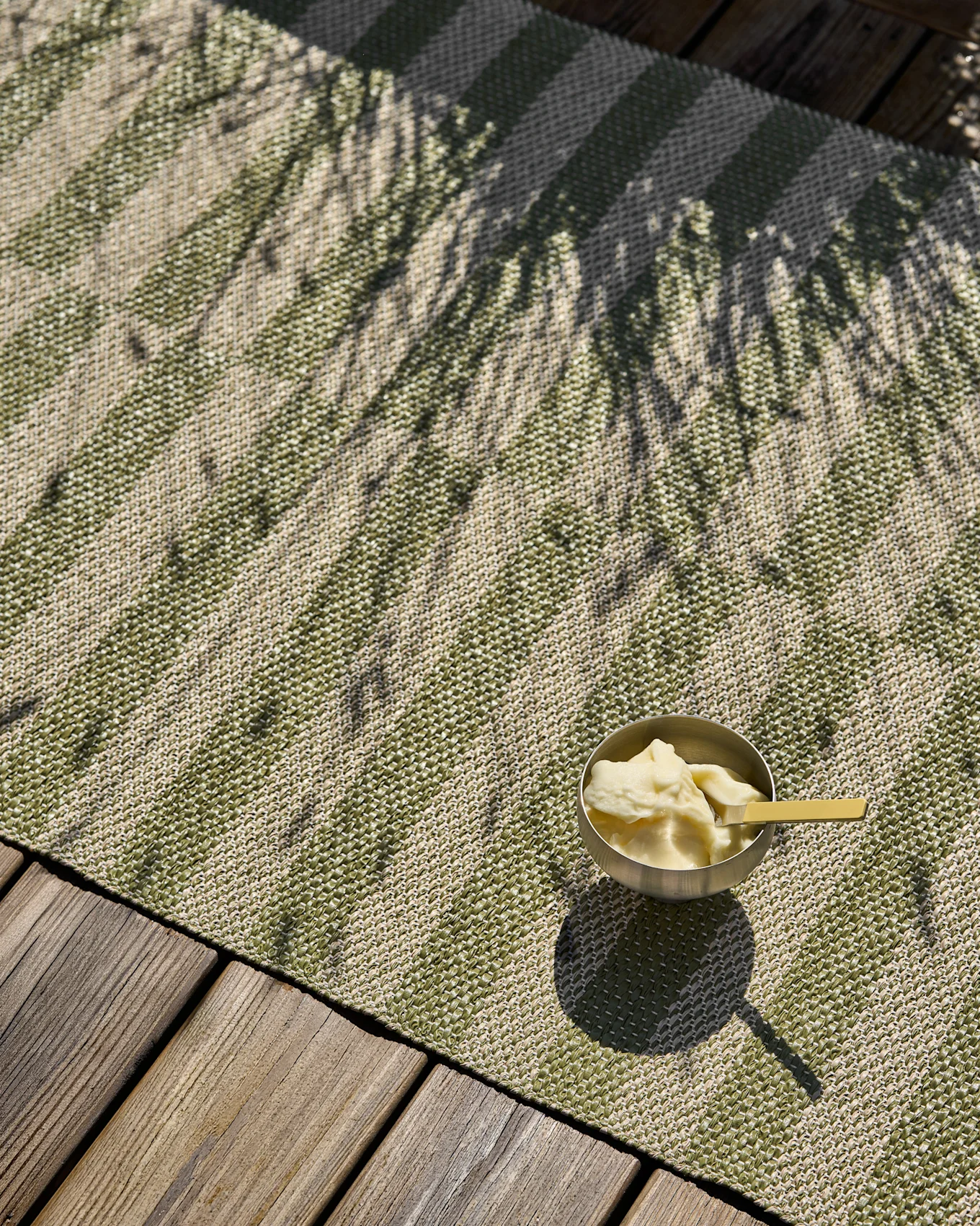 Tapis tissé rayé vert et beige avec un bol de glace et des ombres de palmier sur une terrasse en bois.