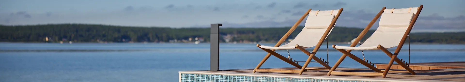 Deux chaises longues en tissu blanc et bois sur une terrasse surplombant un lac avec une forêt au loin.