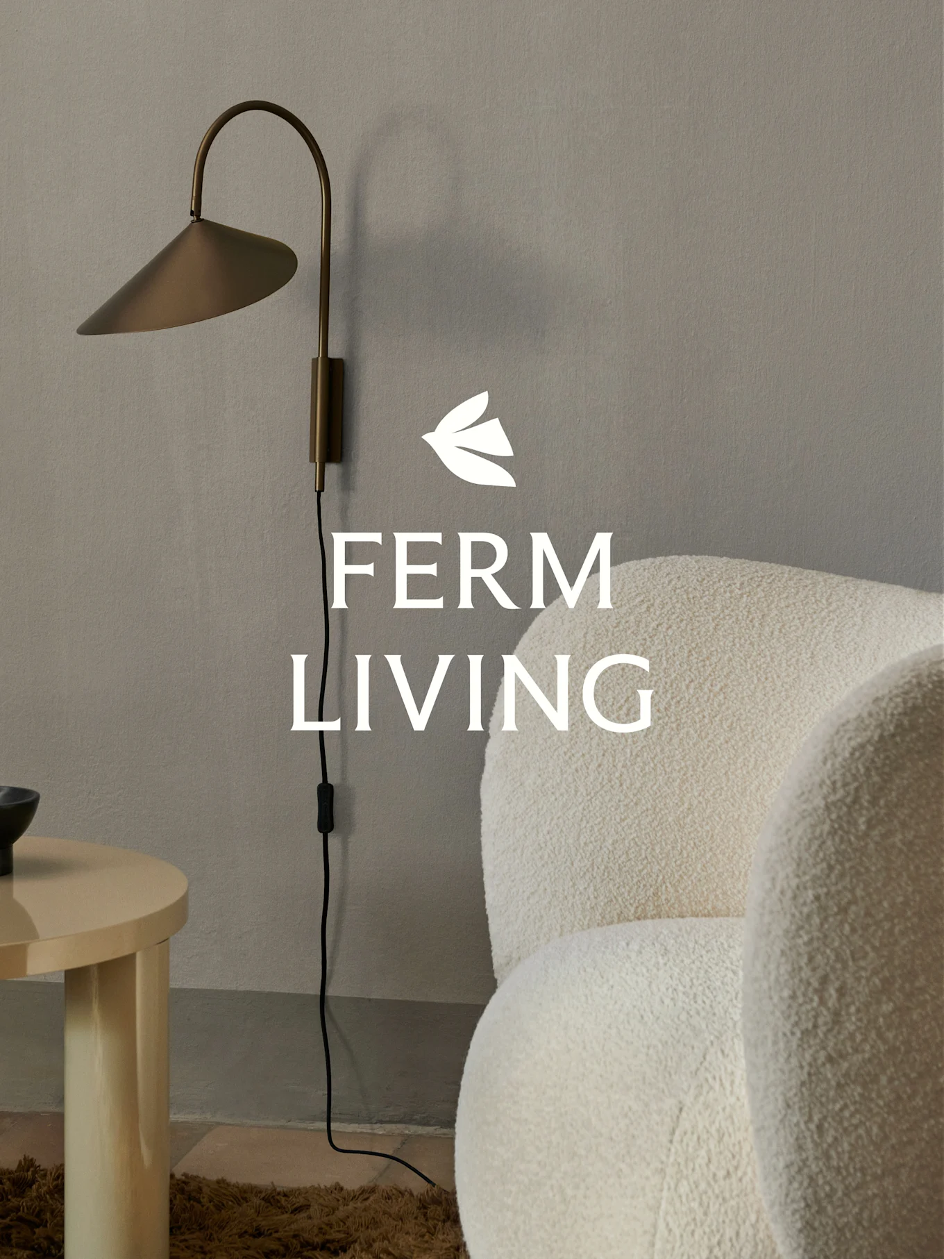 Logo Ferm Living sur une applique murale en laiton, un fauteuil bouclé crème et une table d'appoint en bois clair dans un cadre minimaliste.