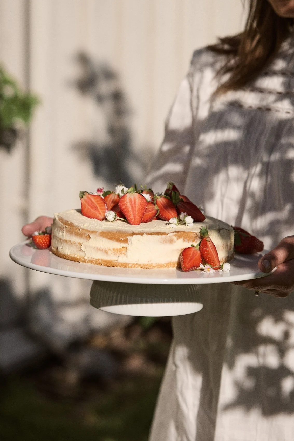 Personne tenant un gâteau rond garni de fraises fraîches coupées en deux et de petites fleurs blanches sur un support à gâteau blanc en extérieur.