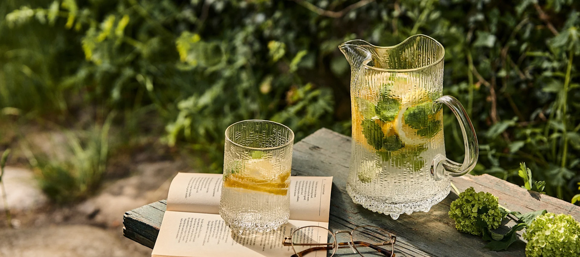 Carafe et verre en verre texturé avec de l'eau citronnée et mentholée, un livre ouvert et des lunettes sur une surface en bois à l'extérieur.