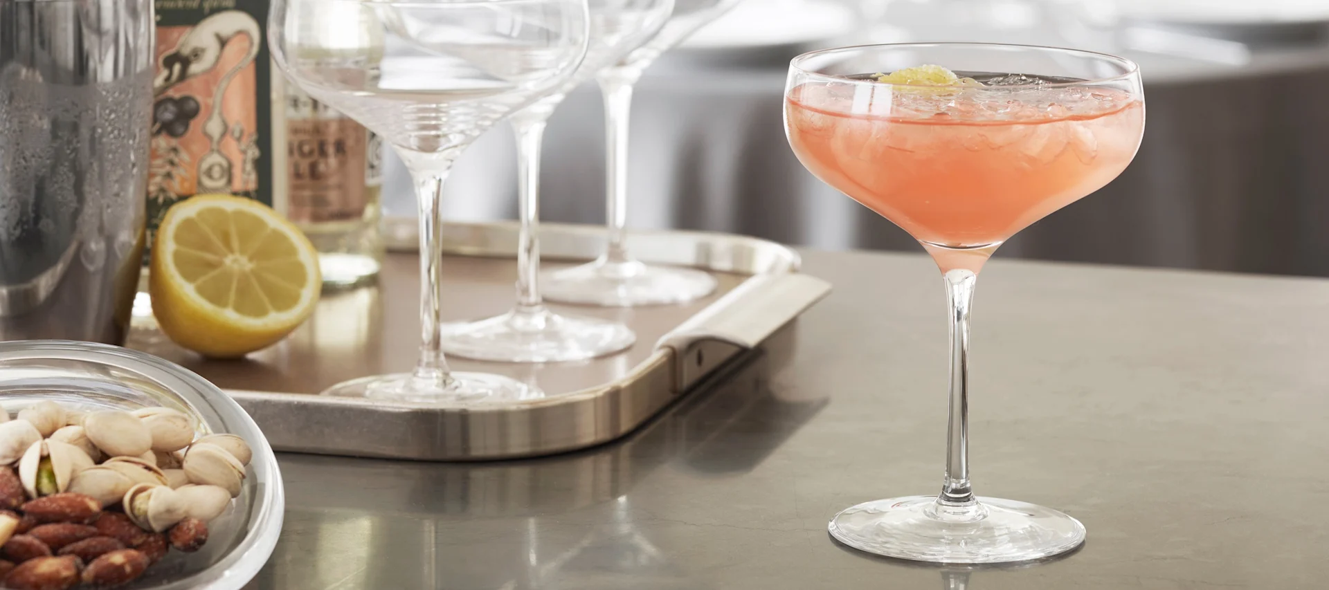 Un cocktail sans alcool à base de jus de pamplemousse servi dans un verre à cocktail Holmegaard Cabernet est posé sur un comptoir de cuisine, avec des ustensiles à cocktail en arrière-plan.