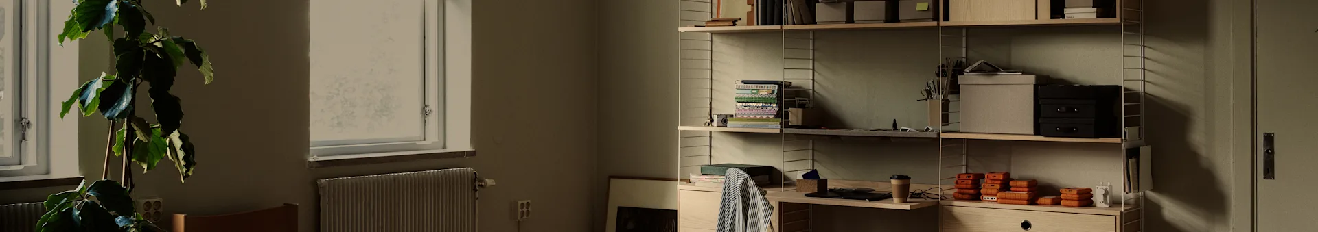 Un bureau à domicile soigné avec une grande plante près d'une fenêtre et une étagère en bois remplie de livres, de boîtes et de disques durs portables oranges.