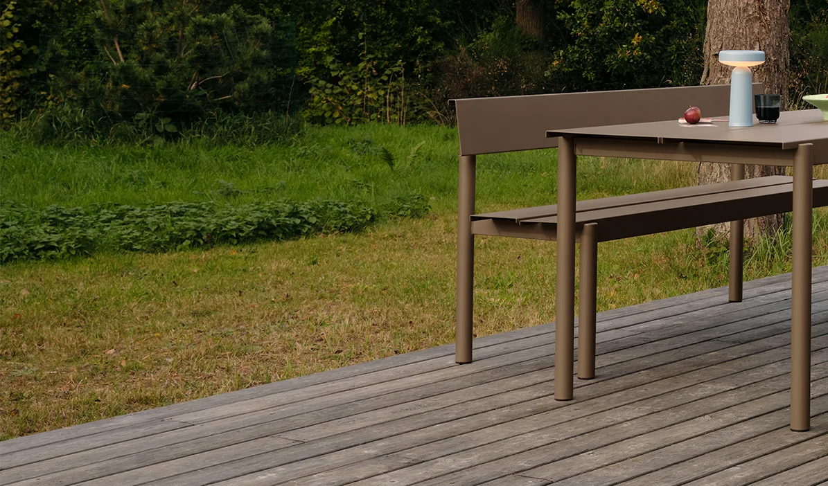 Table et banc de salle à manger modernes couleur taupe sur une terrasse en bois, avec une lampe portable et des pommes sur la table.