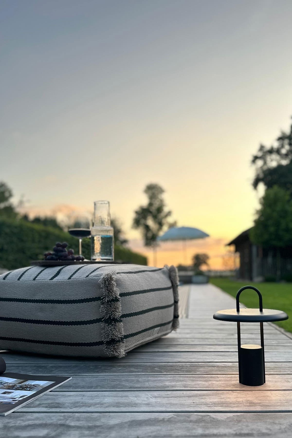 Une terrasse confortable avec un sol en bois, sur lequel repose le coussin d'assise Lina de OYOY, à côté d'une lampe de table Pier noire de Stelton.