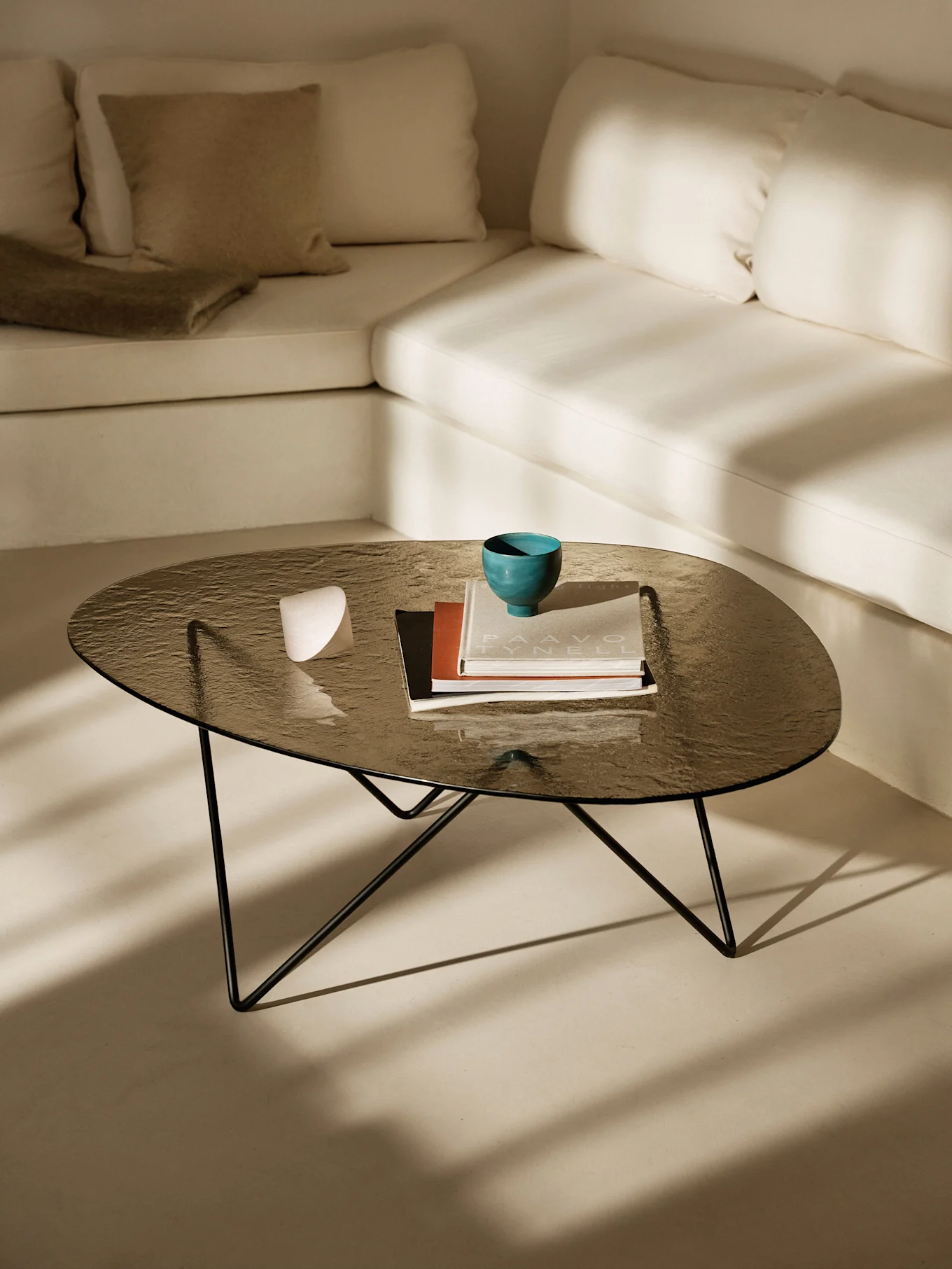Table basse en verre foncé, bol sarcelle, livres, objet blanc. Canapé clair en L, ombres du soleil.
