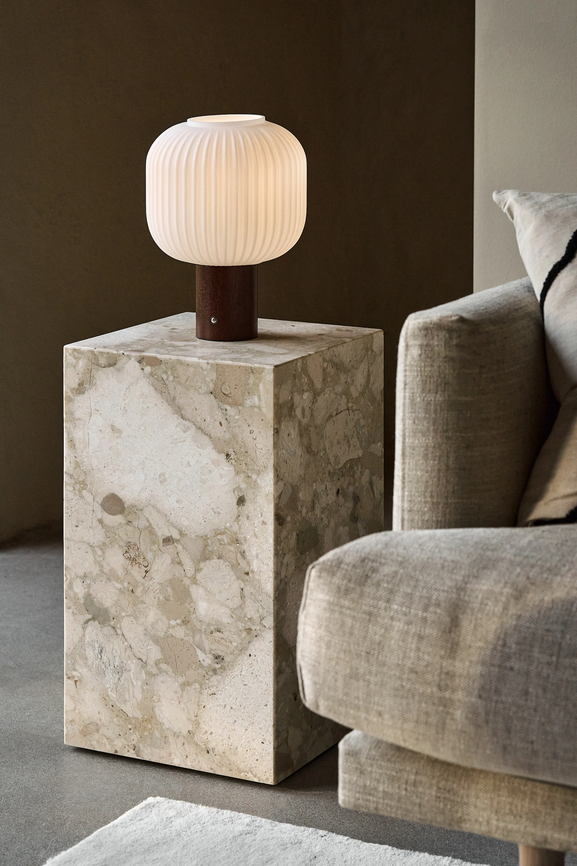 Une lampe de table blanche striée et allumée avec une base en bois sur une table d'appoint en marbre beige à côté d'un canapé clair.