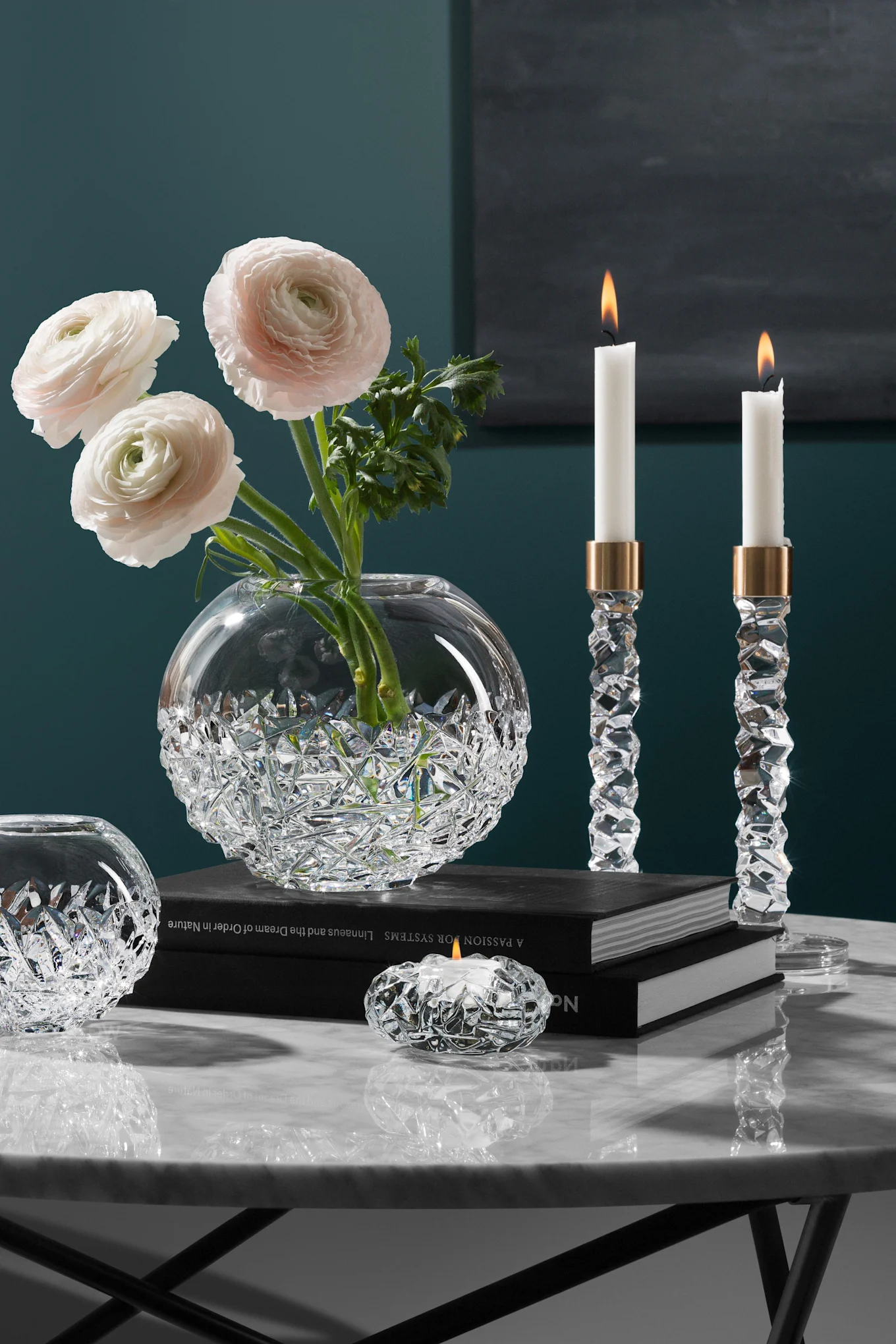 Vase en cristal élégant avec des renoncules roses, deux bougeoirs en cristal allumés et un porte-bougie sur une table en marbre.
